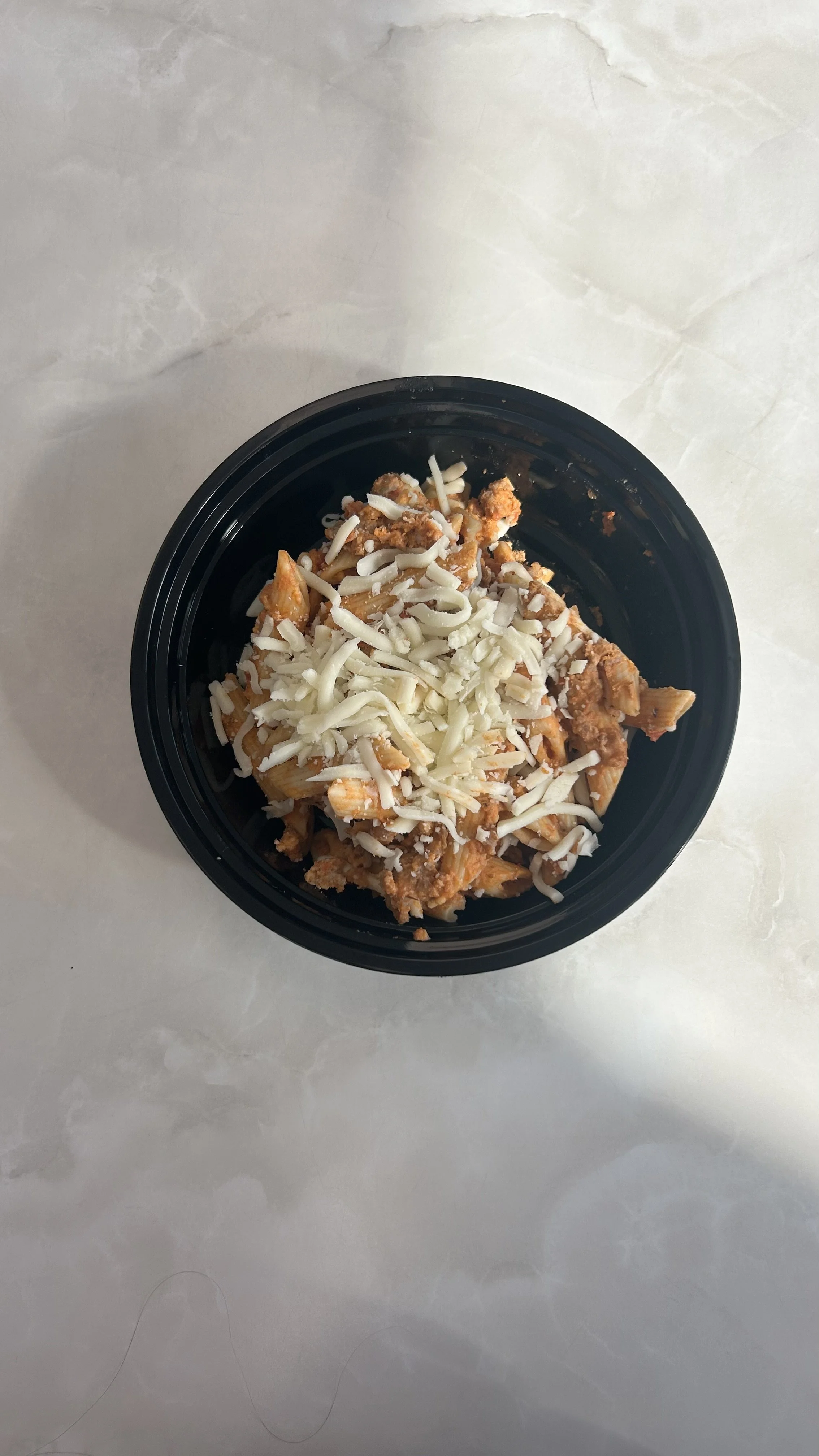A black bowl of pasta with shredded cheese on top, placed on a marble surface.