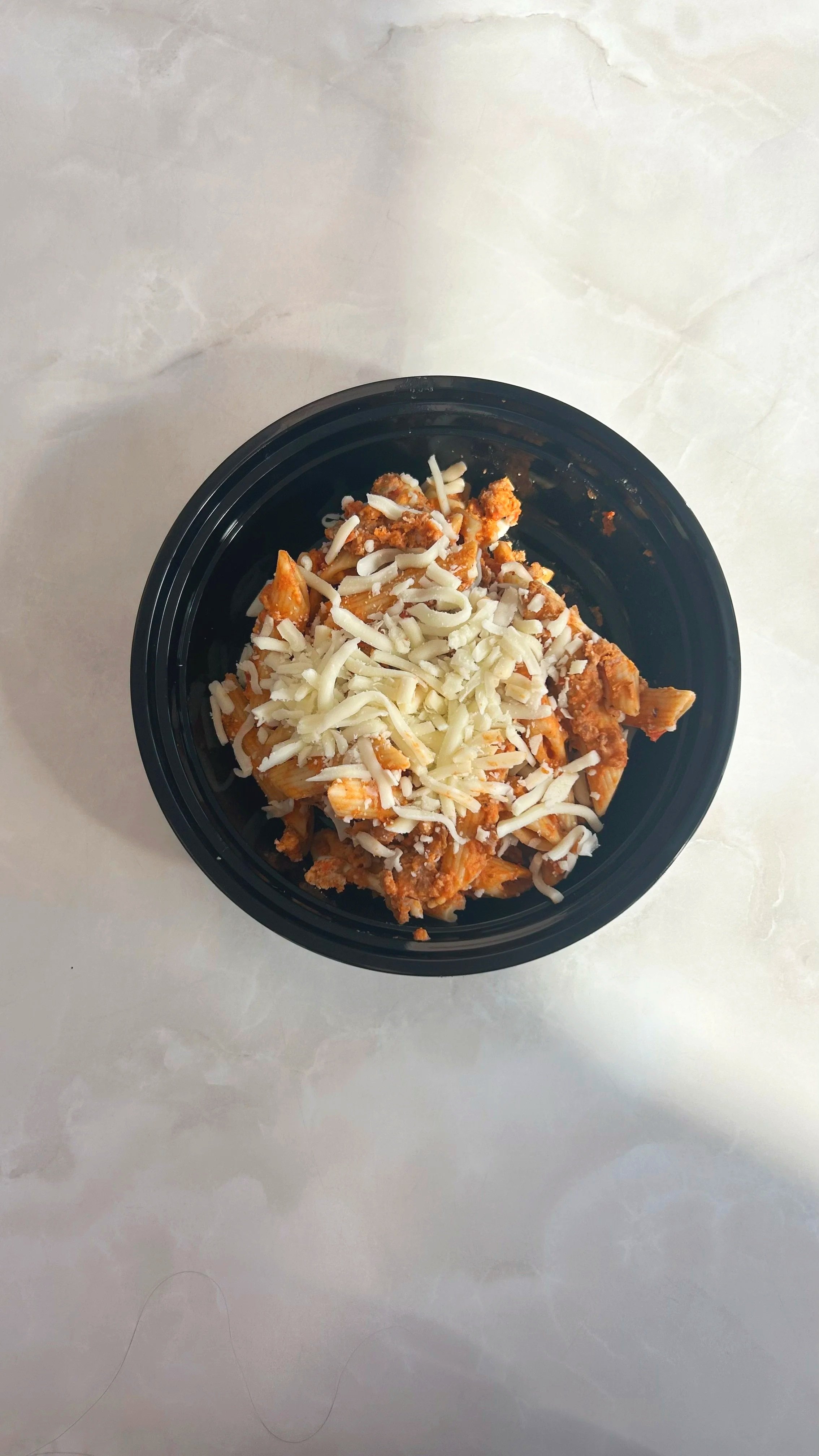 A black bowl of pasta with shredded cheese on top, placed on a white marble surface.