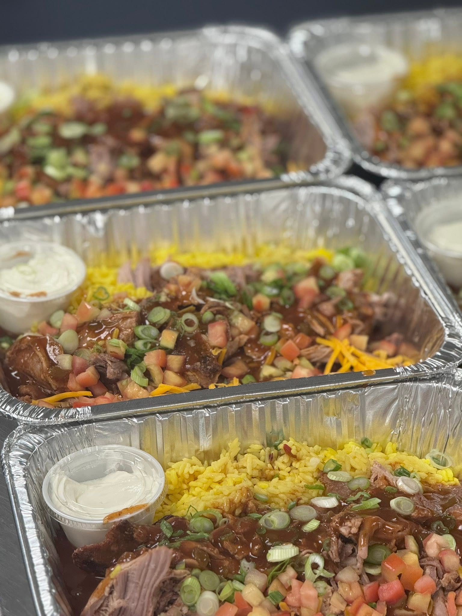 Multiple aluminum takeout containers of food, including shredded yellow rice, meat with gravy, diced vegetables, salad, and small containers of sauces or dips.
