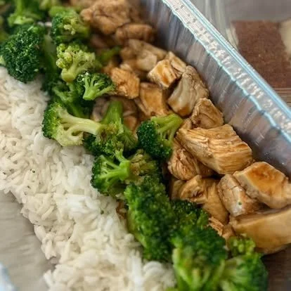 Cooked broccoli florets, white rice, and chunks of cooked chicken inside a foil food container.