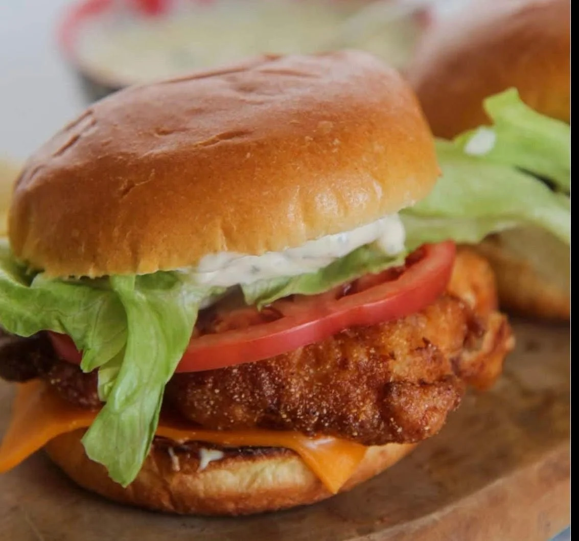 A close-up of a chicken sandwich with lettuce, tomato, cheddar cheese, and a fried chicken fillet in a bun.