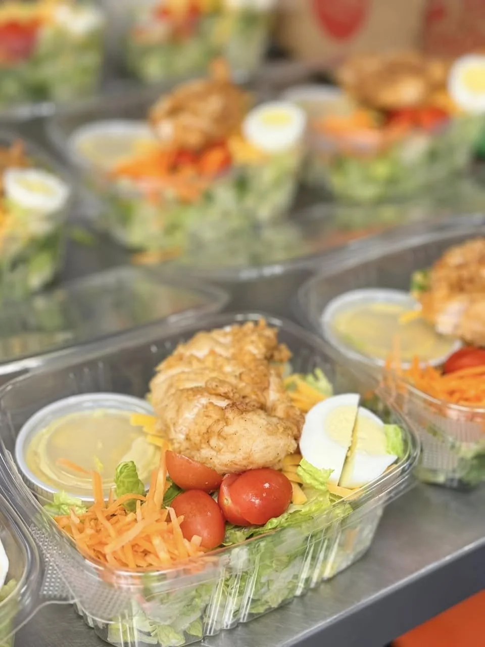 Pre-packaged salad with fried chicken, cherry tomatoes, shredded carrots, hard-boiled eggs, and salad dressing in small containers.