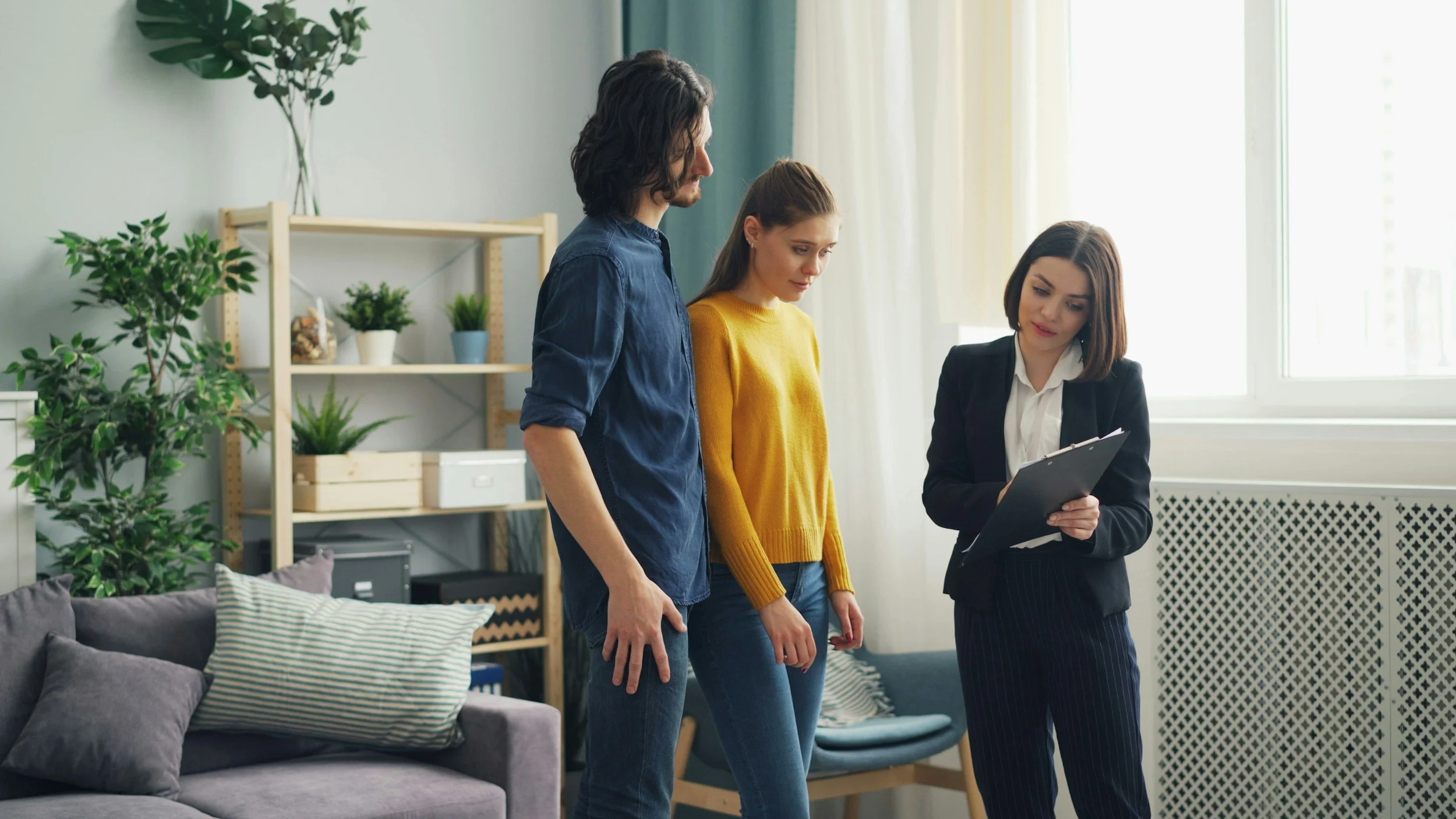 Three women in an office, two standing and looking at a clipboard held by the third woman who is wearing a business suit, with plants and office furniture in the background.