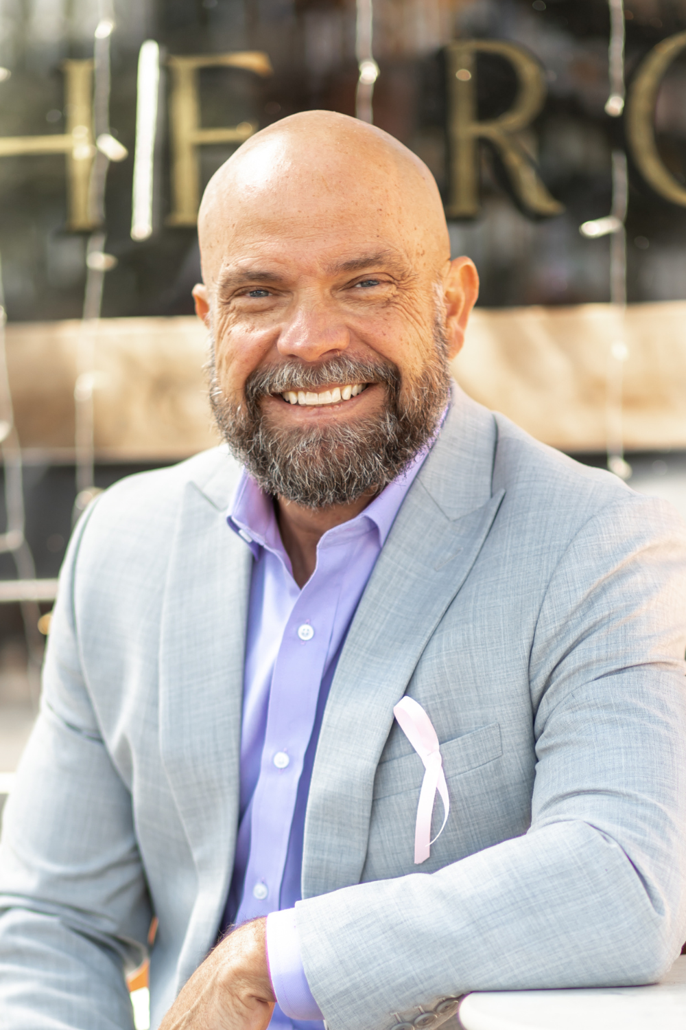 Smiling man with a bald head and beard wearing a light gray suit jacket and purple dress shirt, sitting outdoors with a blurred background of a building and Windows.