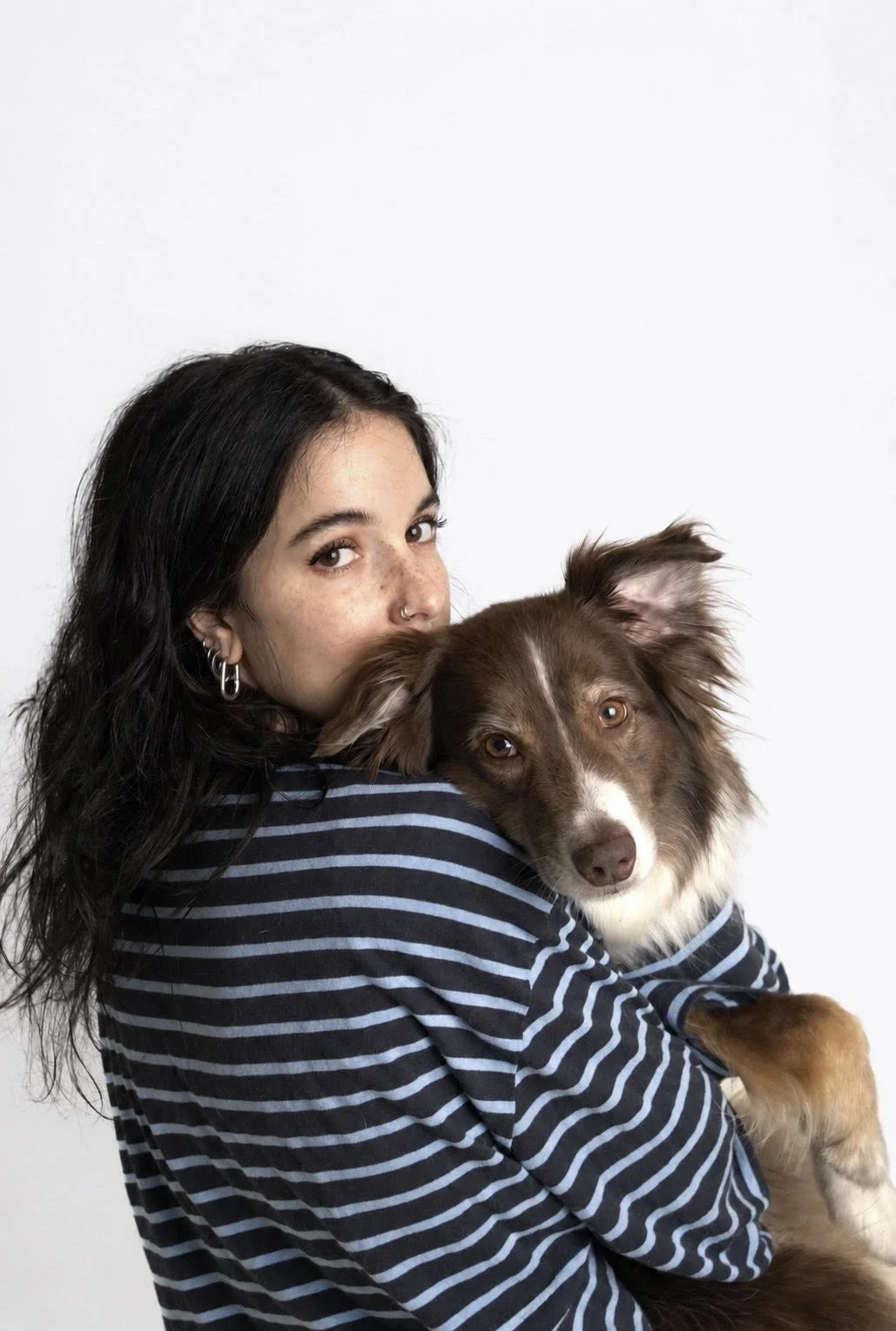 Une jeune femme aux cheveux noirs, portant un pull à rayures bleues et blanches, tient un chien marron et blanc dans ses bras.