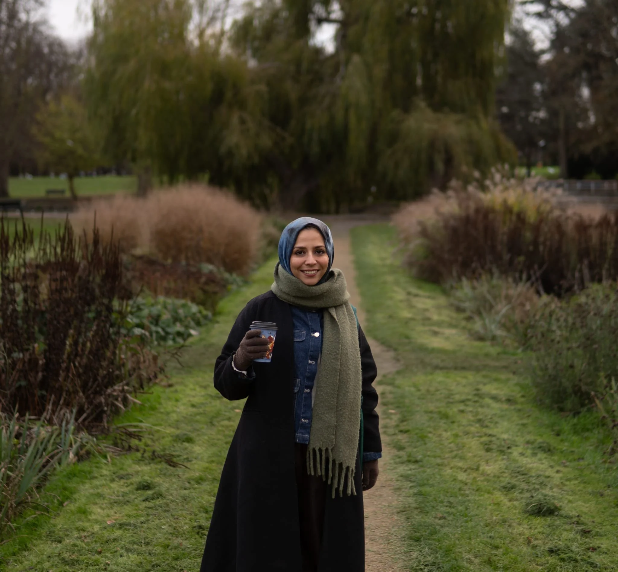 A woman wearing a headscarf and coat standing outdoors in a park or garden, smiling and holding a travel mug, with a pathway and trees in the background.