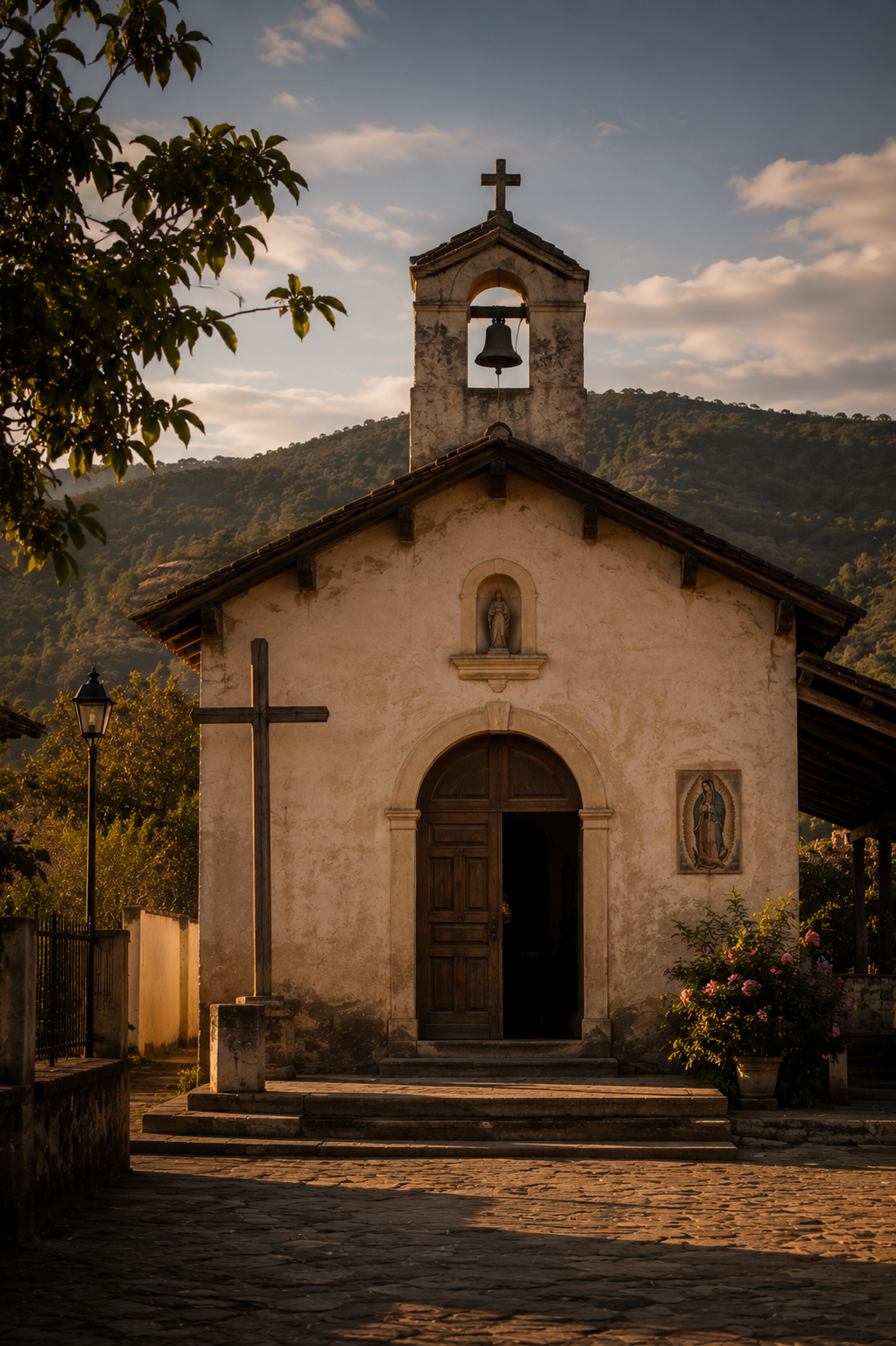A small, rustic church with a wooden door, a cross on the side, and religious statues on the front facade, set against a backdrop of green hills and a partly cloudy sky.