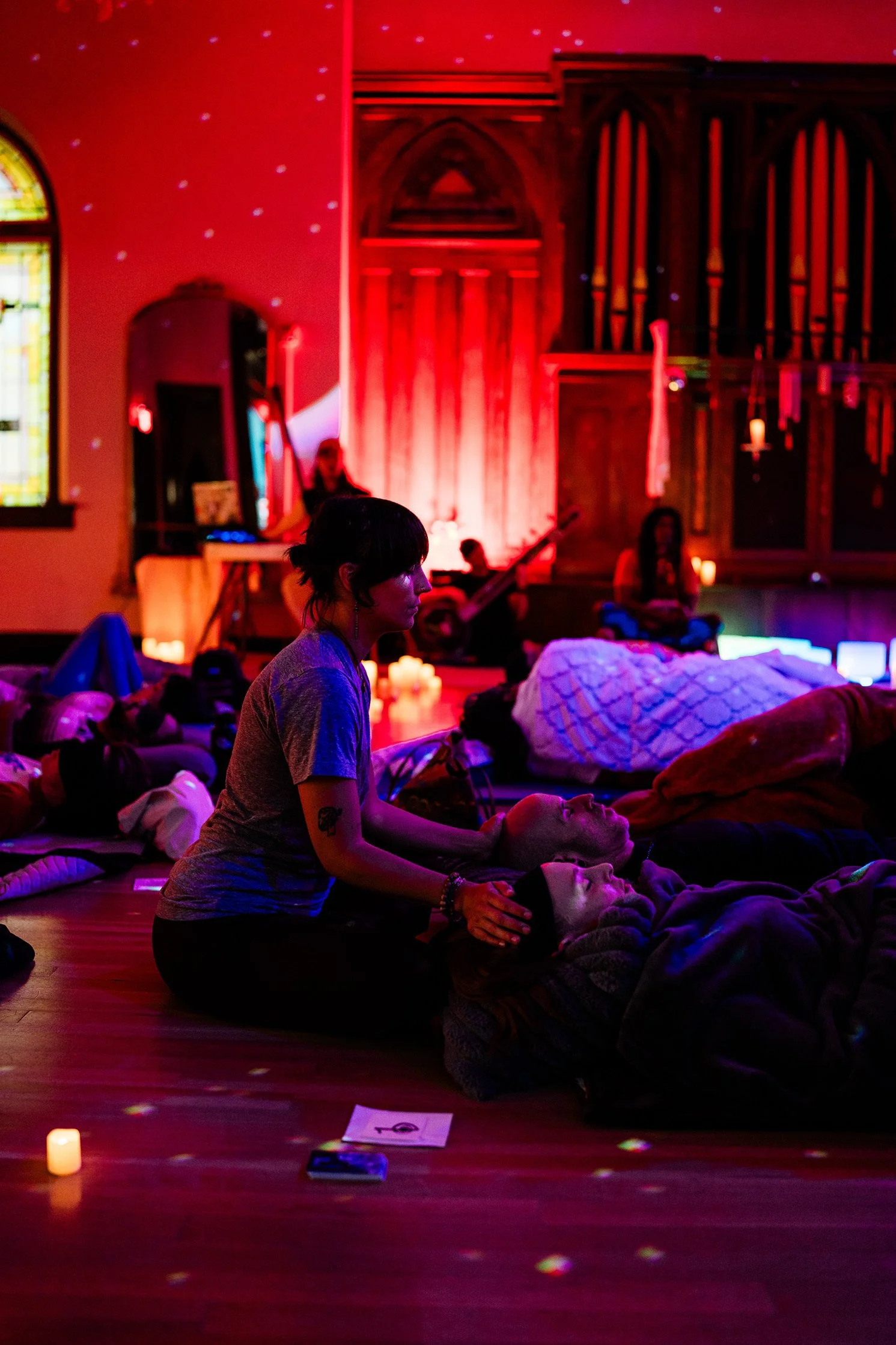 A group of people participating in a meditation or Reiki session in a dimly lit room with red and purple lighting, candles, and projections of light on the wall.