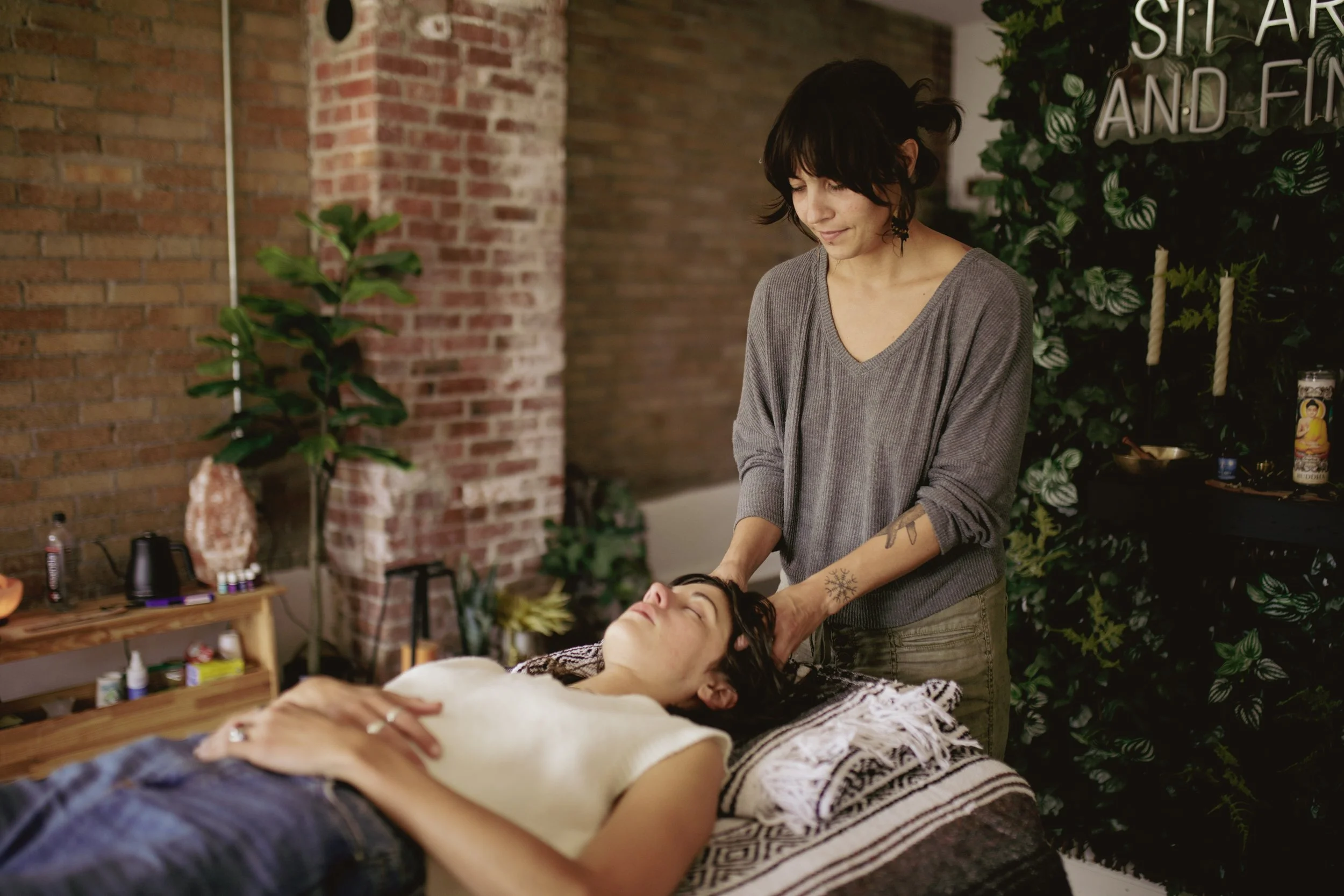 A woman giving a massage to another woman lying on a massage table in a cozy, decorated massage room with brick walls, plants, and a sign that reads 'Star and Fina'.