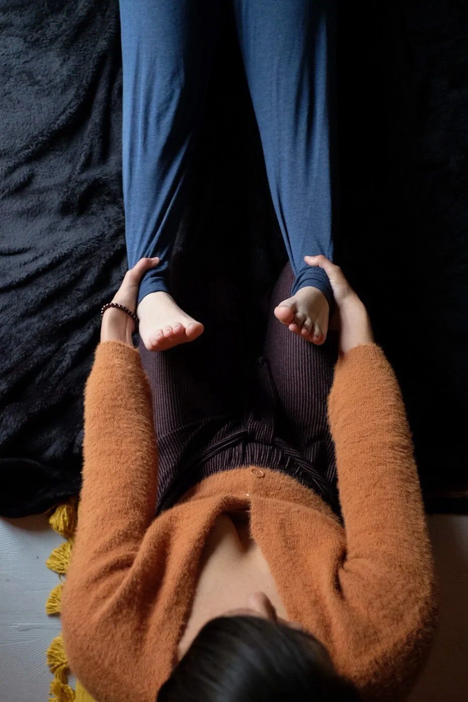 Person lying on a black fuzzy blanket, wearing a rust-colored sweater and brown striped pants, holding a child by the ankles, who is lying above them, wearing blue pants.