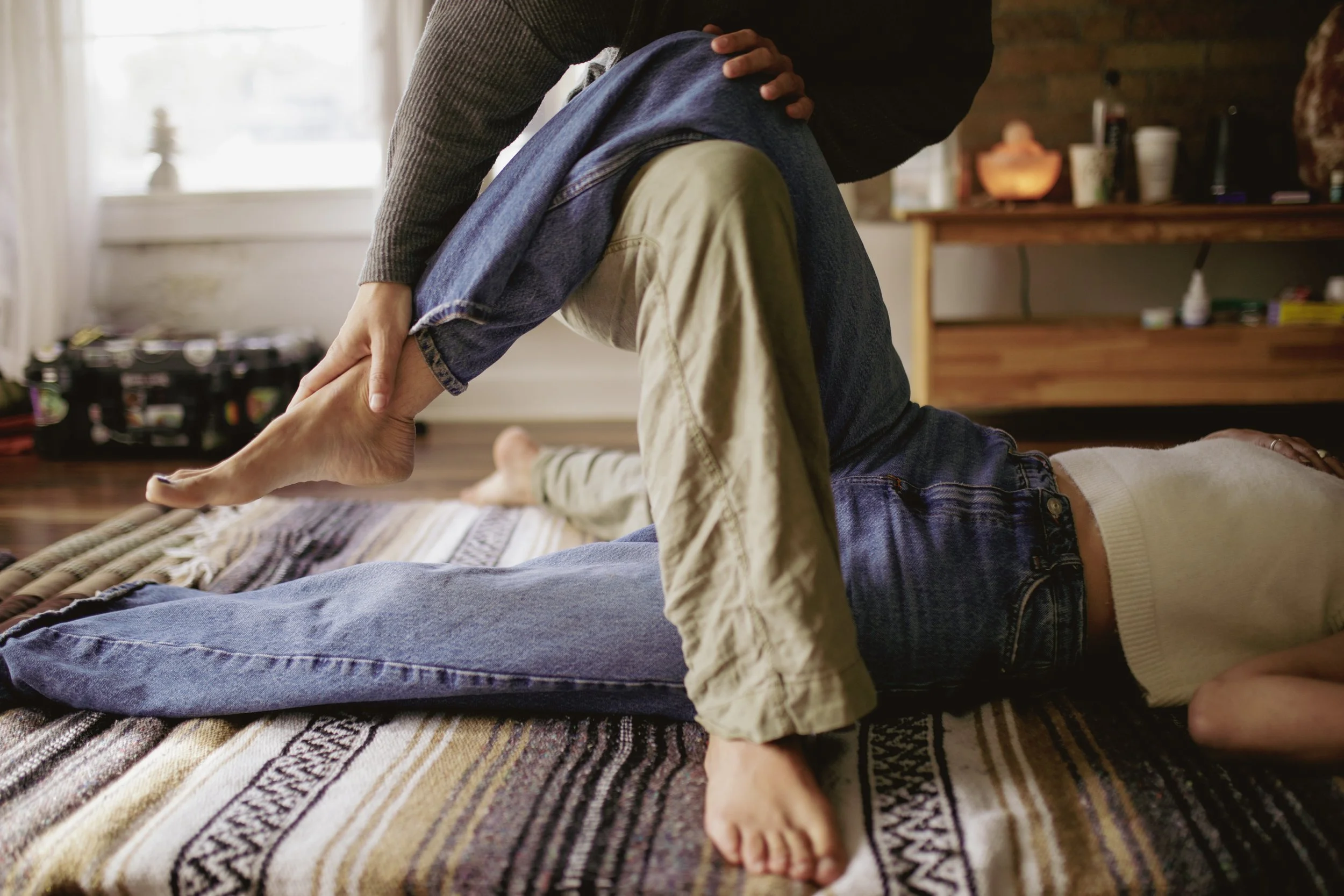 A person in a gray sweater and jeans performs a yoga pose or stretch with another person lying on a patterned rug, in a cozy room with wooden shelves and a window with natural light.