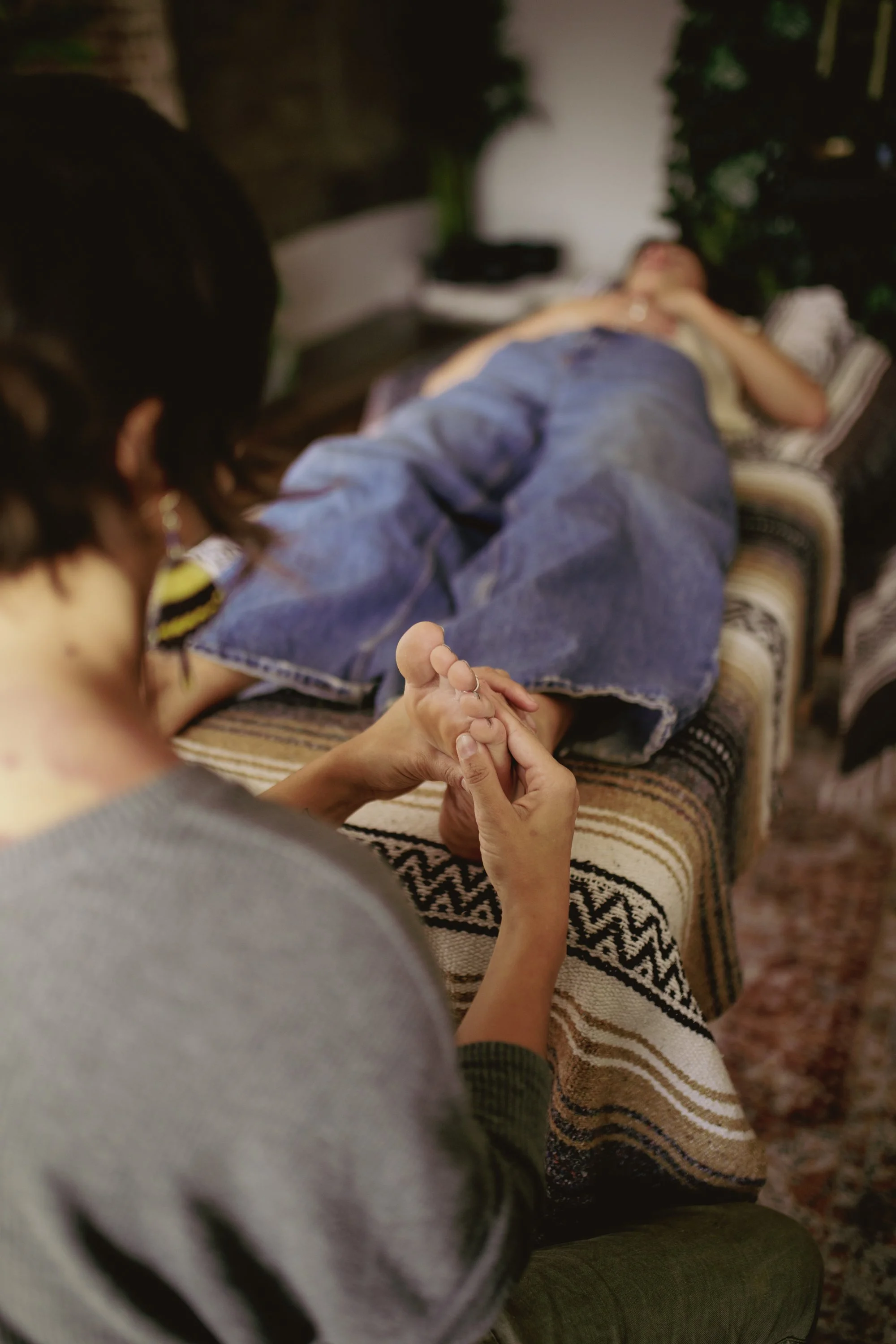 A person lying on a bed with their hand being held by a woman. The person is dressed in a blue shirt and gray pants, lying on their back with eyes closed. The woman has dark hair and is wearing a gray sweater, holding the person's hand gently. The se