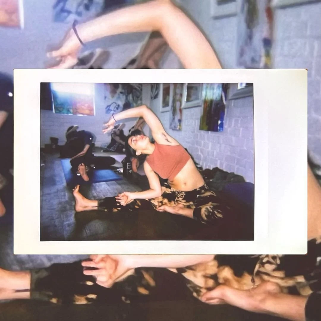 Photo of a woman in a yoga pose on a black mat, with her arms stretched overhead, smiling, wearing an orange tank top and patterned pants. In the background, a person is sitting on a mat in a room with white brick walls and artwork displayed.