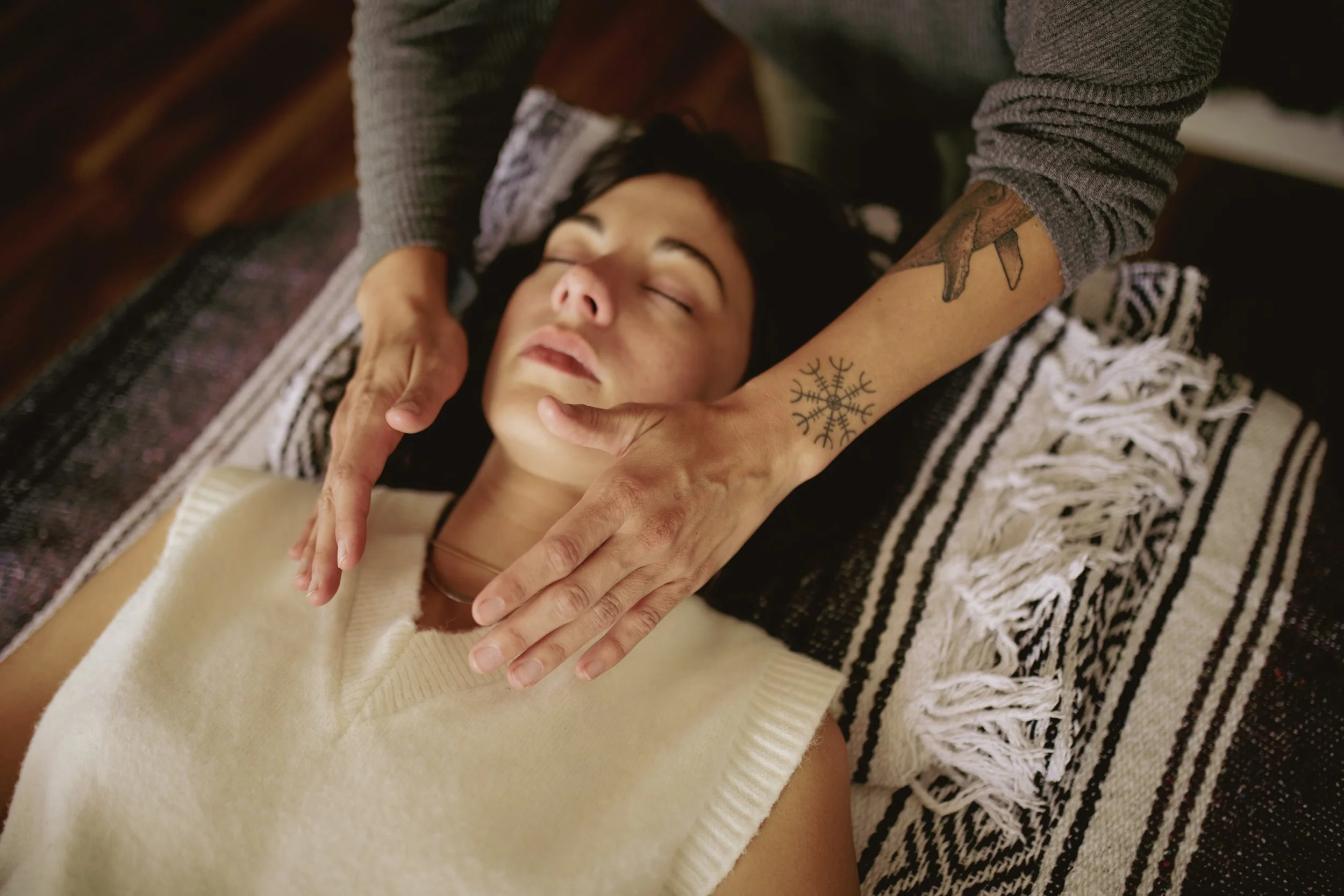 A woman receives a facial massage from a person with tattoos on their arm. The woman is lying down with her eyes closed, wearing a cream-colored sweater, while the person with tattoos wears a gray long-sleeve shirt.