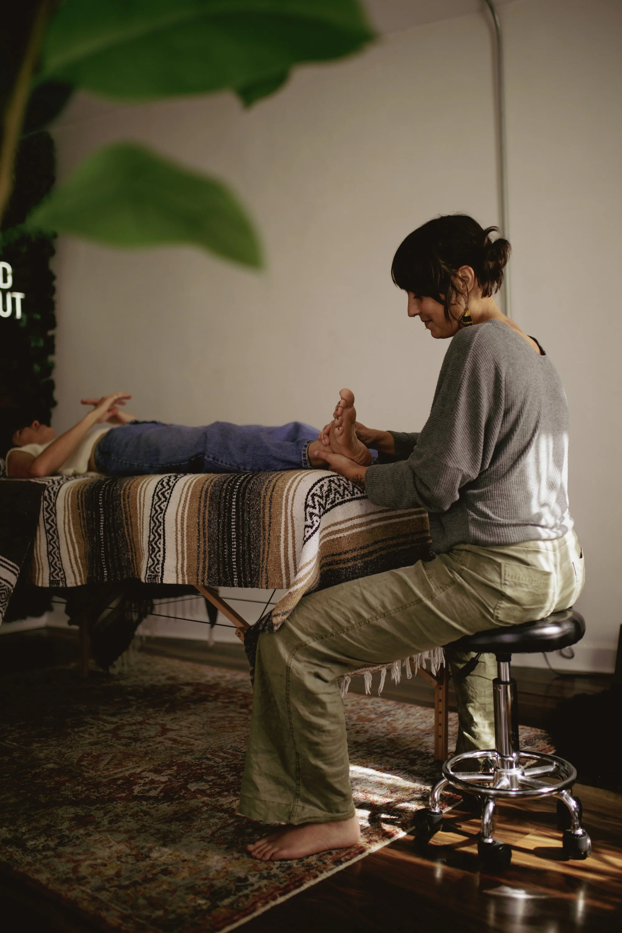 A woman receives a foot massage from a therapist in a cozy room with a patterned rug, a massage table covered with a woven blanket, and a large leafy plant in the background.