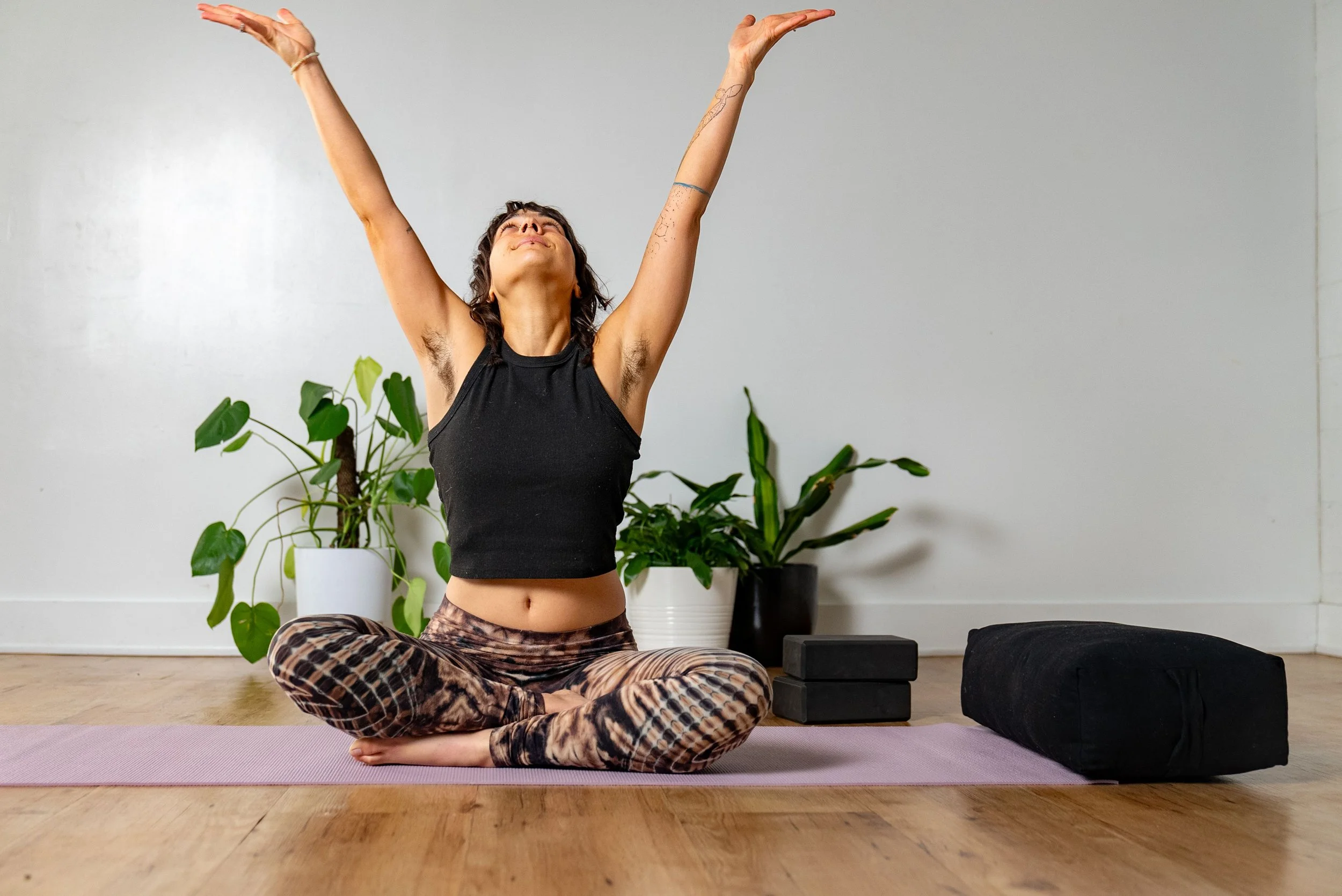 A woman practicing yoga indoors, sitting cross-legged on a pink yoga mat with her arms raised above her head and eyes closed, surrounded by green potted plants, black meditation cushions, and yoga blocks.