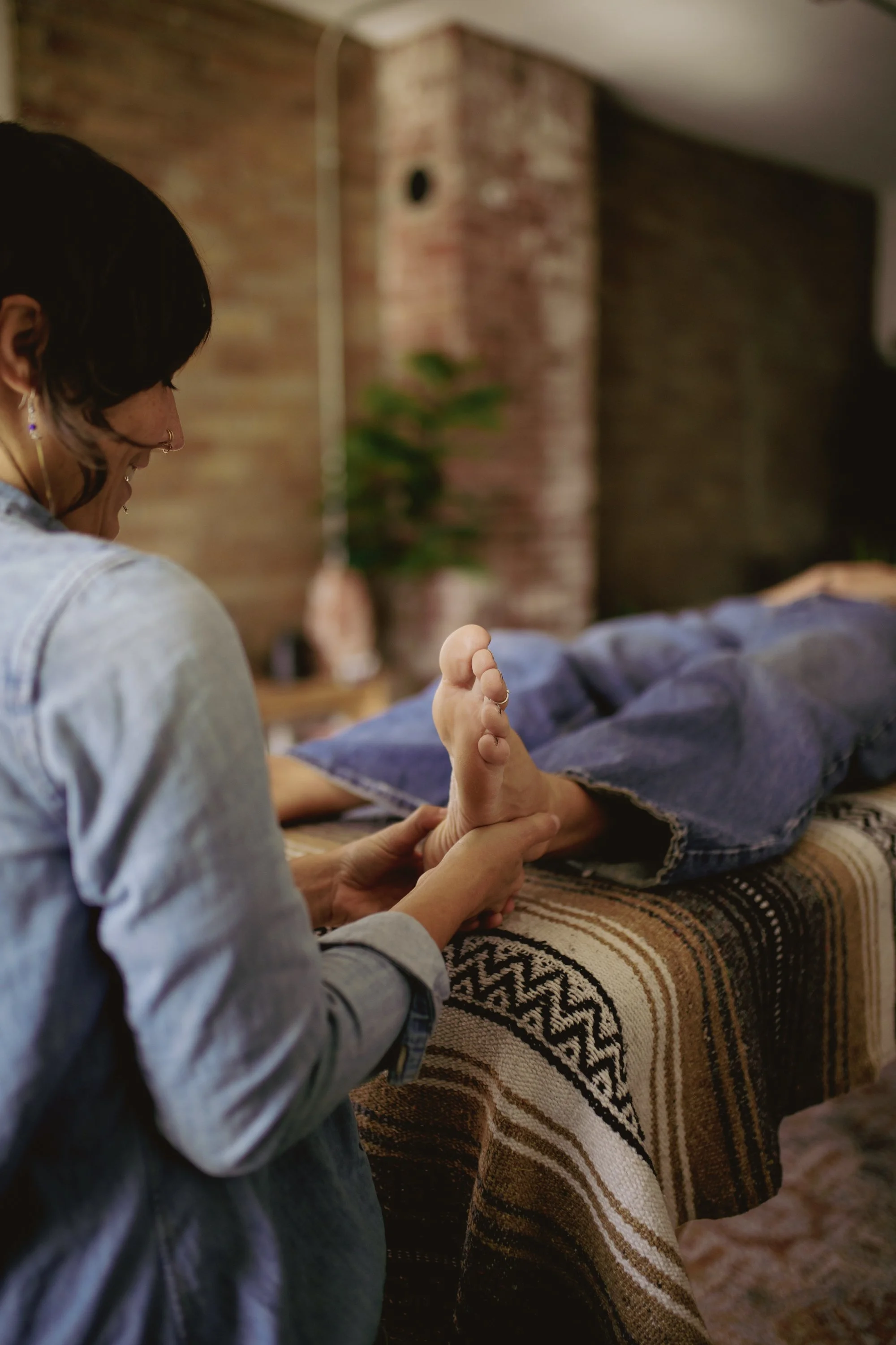 A person with dark hair receiving a foot massage from another person in a cozy indoor setting with a brick wall and plant in the background.