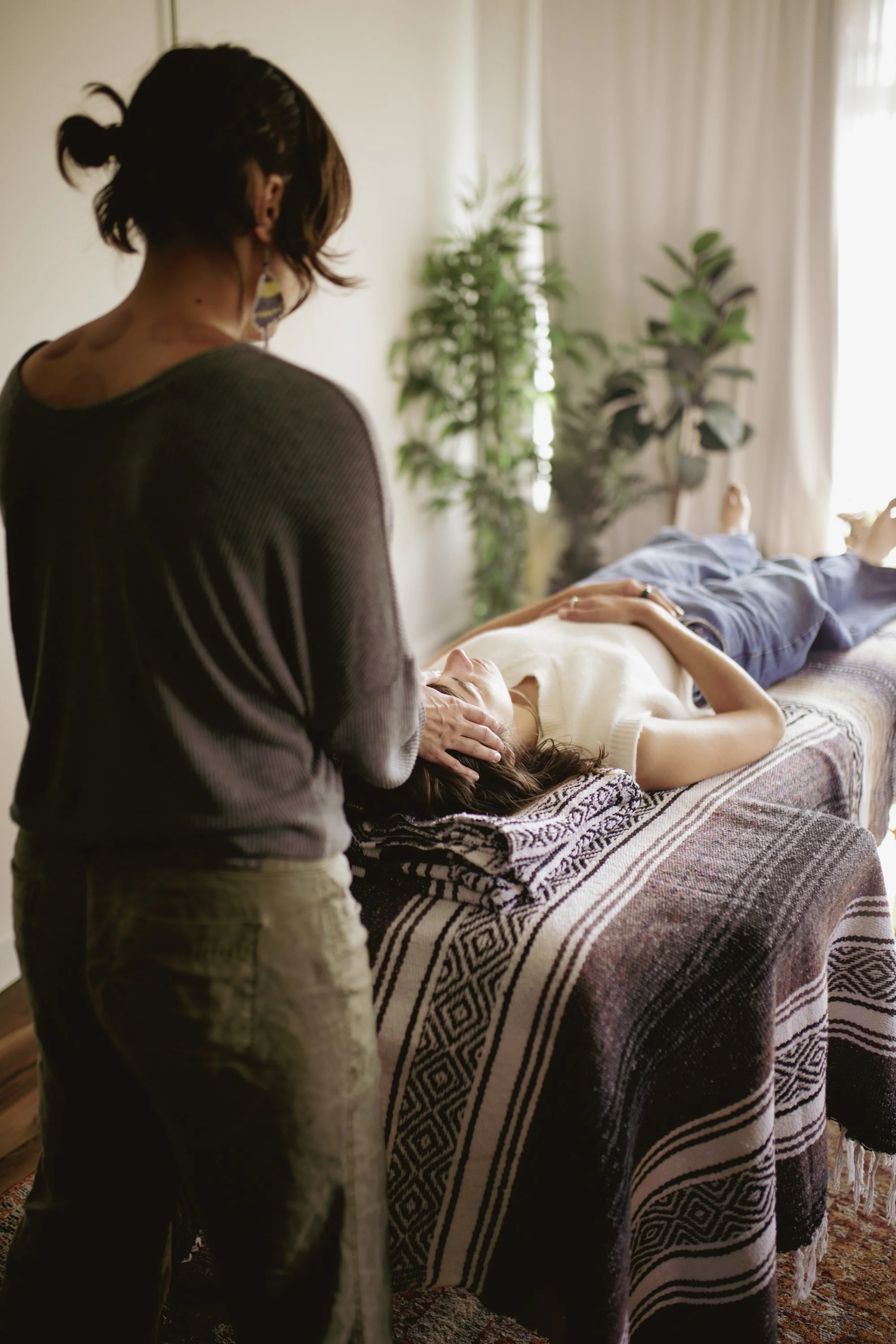 A woman with short dark hair and earrings, seen from behind, is giving a massage or holding the head of a person lying on a bed covered with a black and white patterned blanket. The person lying has long dark hair, and is resting with eyes closed. Th