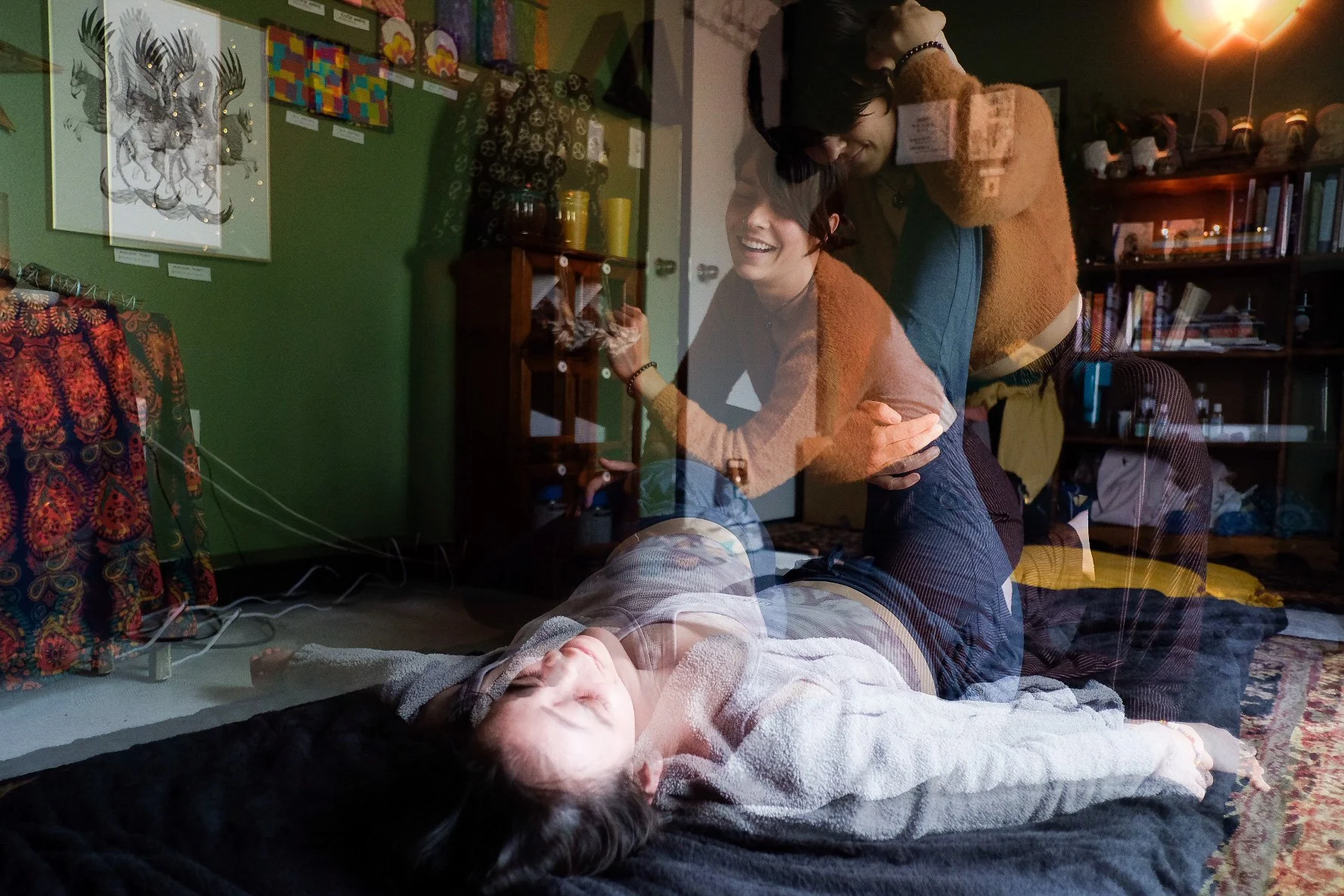 A layered photo shows people practicing restorative yoga or a similar healing practice inside a room with various artwork and books. A person lies relaxed on a blanket with eyes closed, while another supports them, and two others assist or observe ne