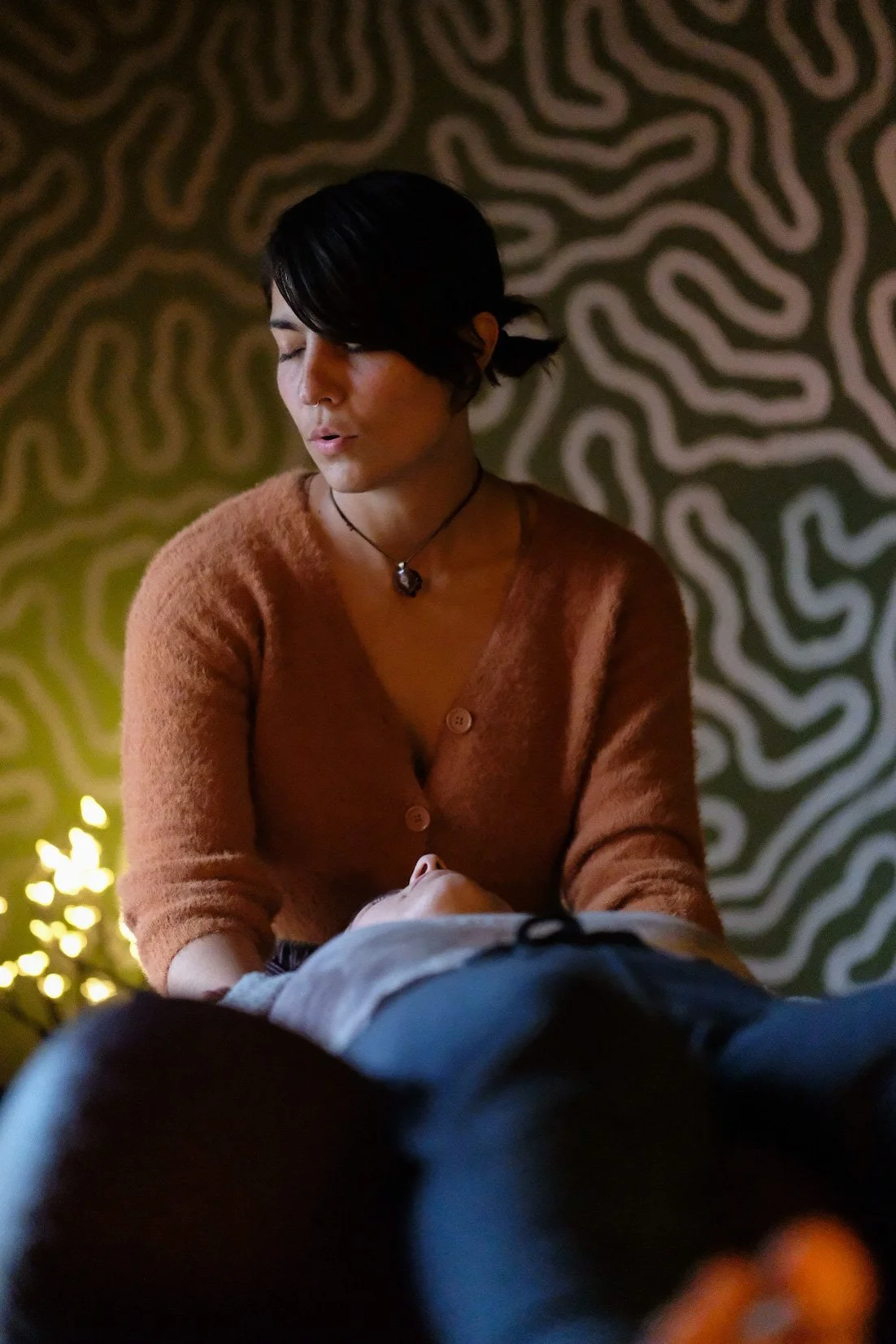 A woman with short dark hair and a necklace, eyes closed, appearing to perform a massage on someone lying down in front of her, in a dimly lit room with a patterned background.
