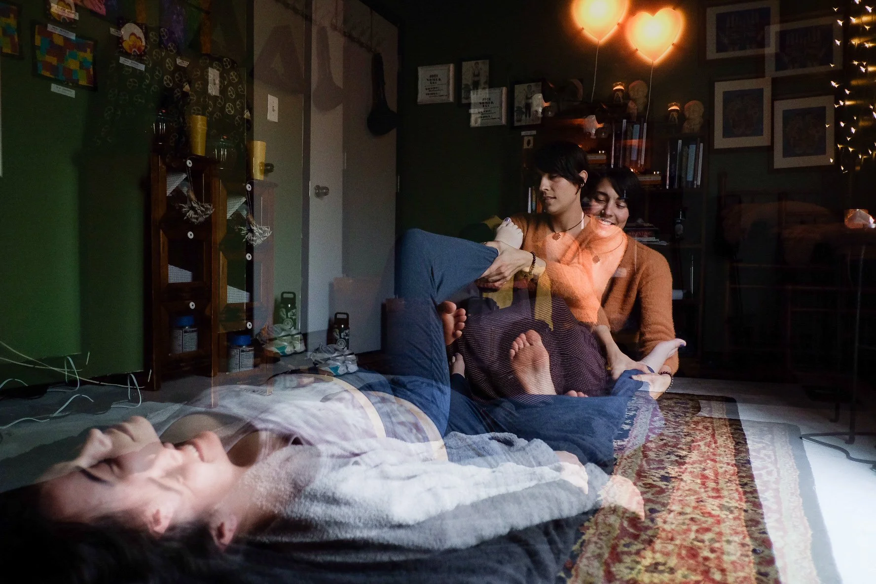 A double exposure photo showing a person lying on the floor and two people sitting on a rug in a living room. The wall is decorated with framed pictures and a bookshelf. Two orange heart-shaped balloons are floating near the ceiling.