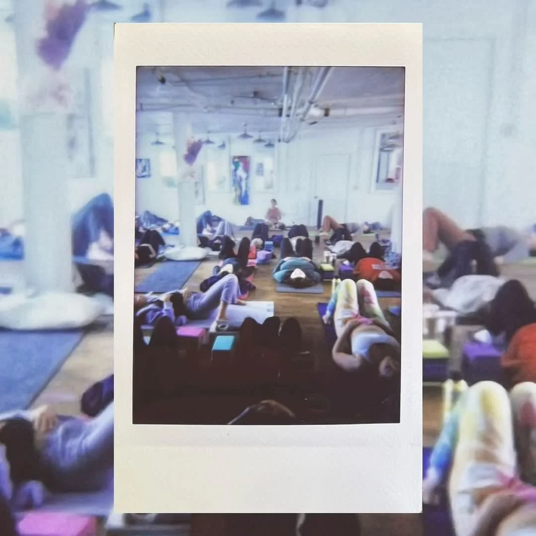 A photo of a yoga or meditation class with people lying on mats in a spacious, well-lit studio. The photo is framed as a print or instant photo placed in front of the scene.