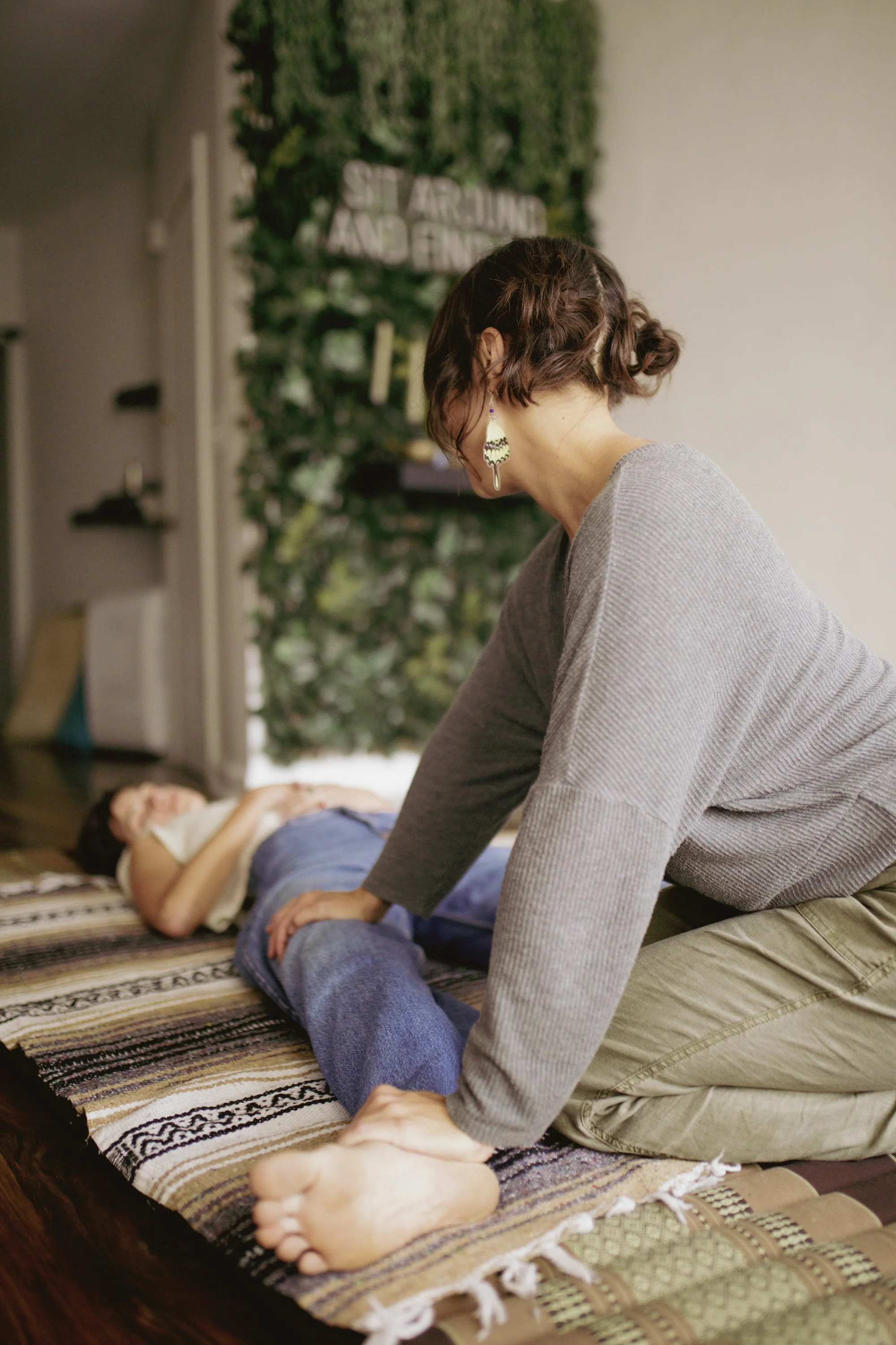A woman receiving a massage from another woman in a cozy room with green plants and a sign that says 'AROUND AND ENJOY'.
