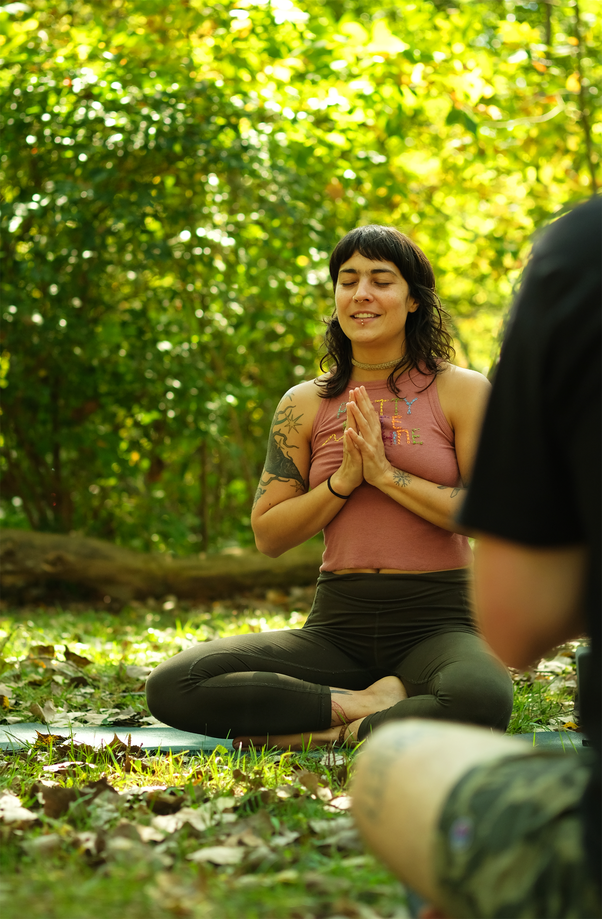 A young woman practicing yoga outdoors in a forest, sitting cross-legged on a mat with her hands in a prayer position, eyes closed, and smiling peacefully.
