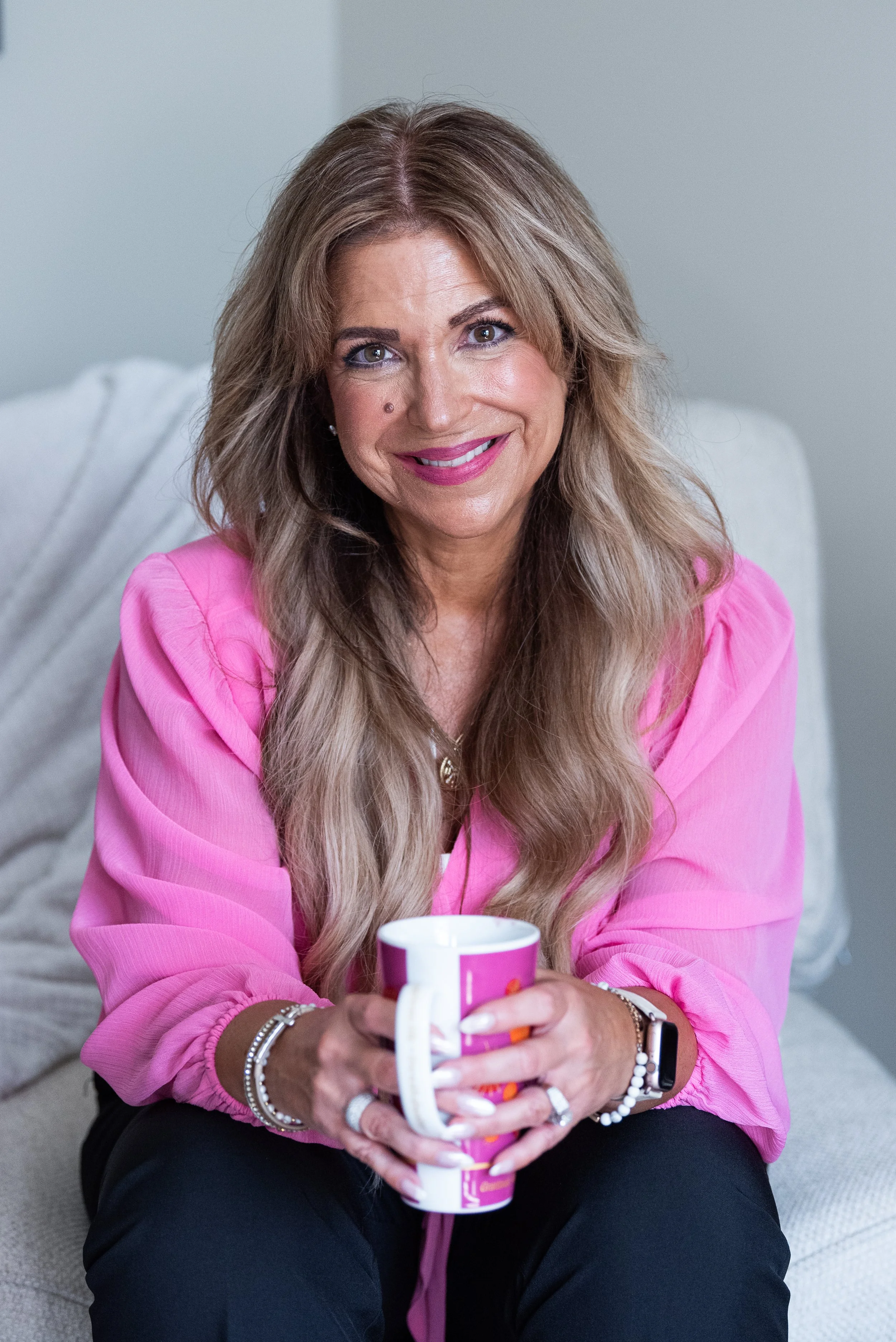 A woman with long wavy blonde hair sitting on a beige couch, wearing a pink long-sleeved shirt, holding a pink and white mug with a beverage. She is smiling, wearing jewelry, and looking at the camera in a cozy indoor setting.