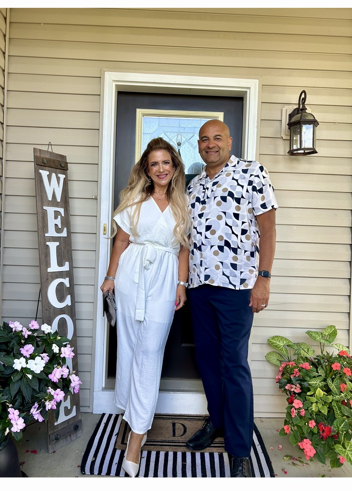 A man and woman standing on a porch in front of a door, smiling at the camera. The woman is wearing a white jumpsuit and holding a purse, the man is wearing a patterned short-sleeve shirt and dark pants. There is a welcome sign on the left, and potted flowers on either side of the doorway.