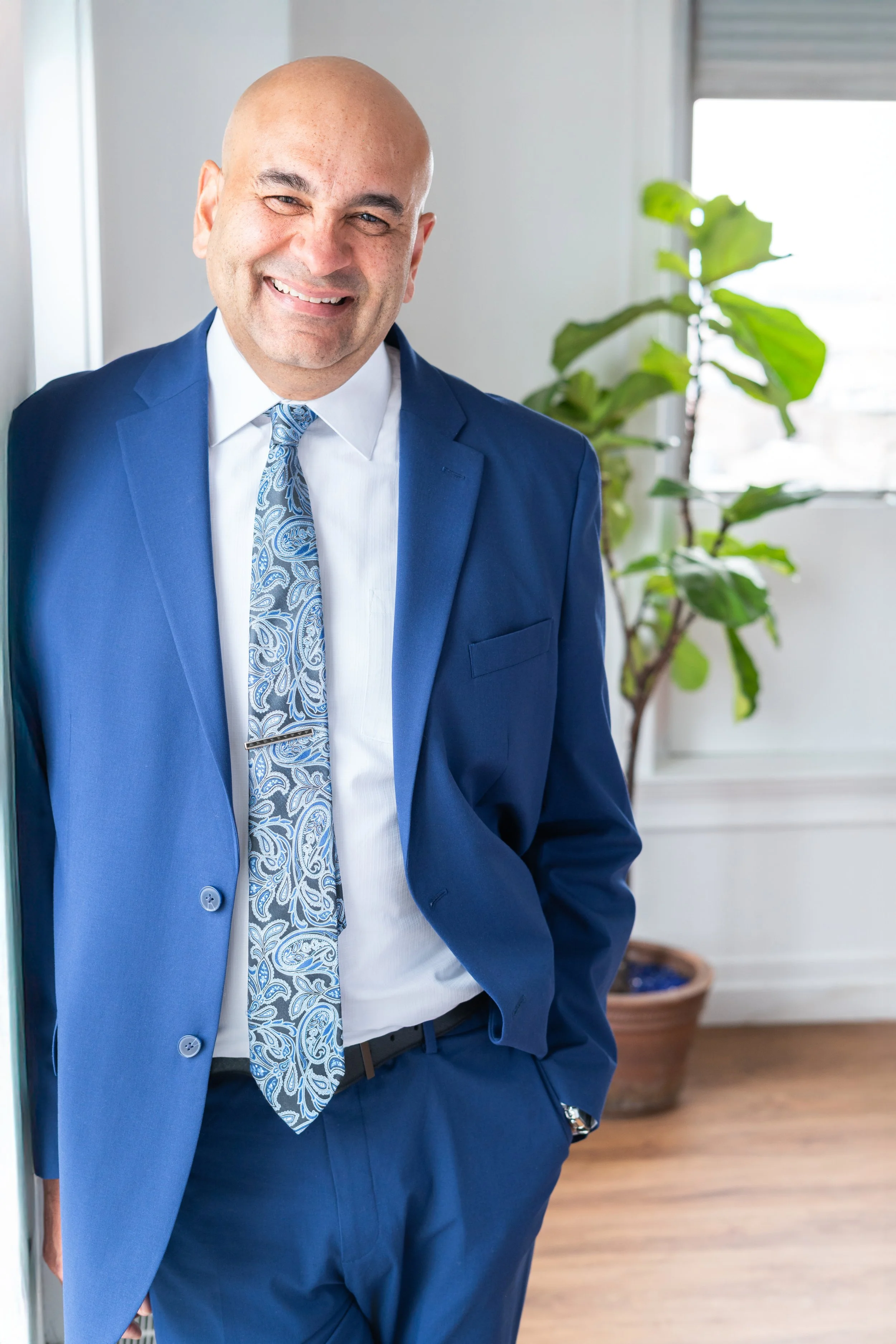 Smiling man in a blue suit with a patterned tie, standing indoors beside a window and a potted plant.