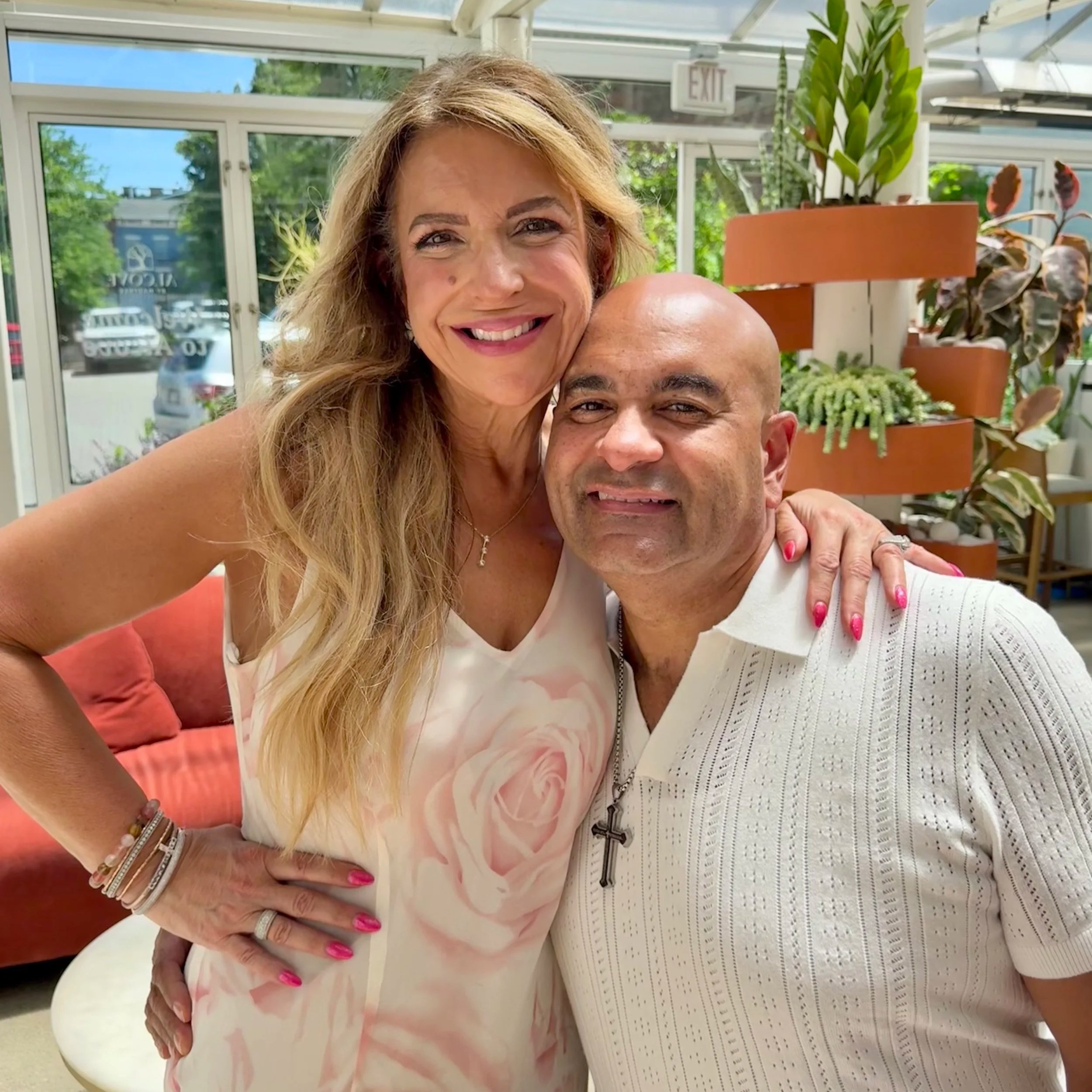 A woman with long blonde hair smiling, wearing a light-colored floral shirt and bracelets, alongside a man with short dark hair in a short-sleeved knit shirt, indoors with glass windows and potted plants behind them.