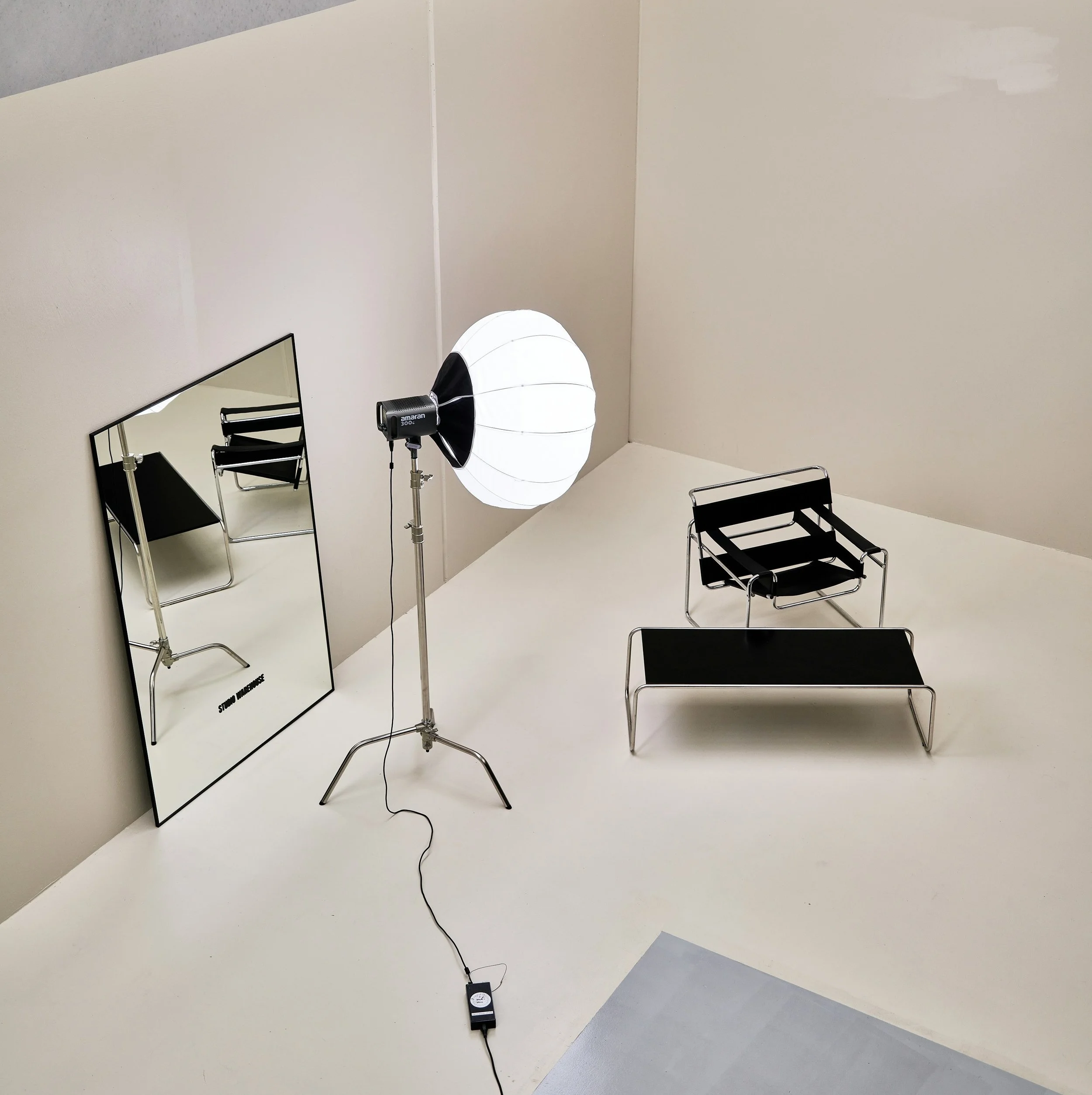 A photography studio setup with a large white umbrella light, a rectangular mirror, three black chairs, and a floor sensor connected by a black cord. The room has plain beige and gray walls and a white floor.