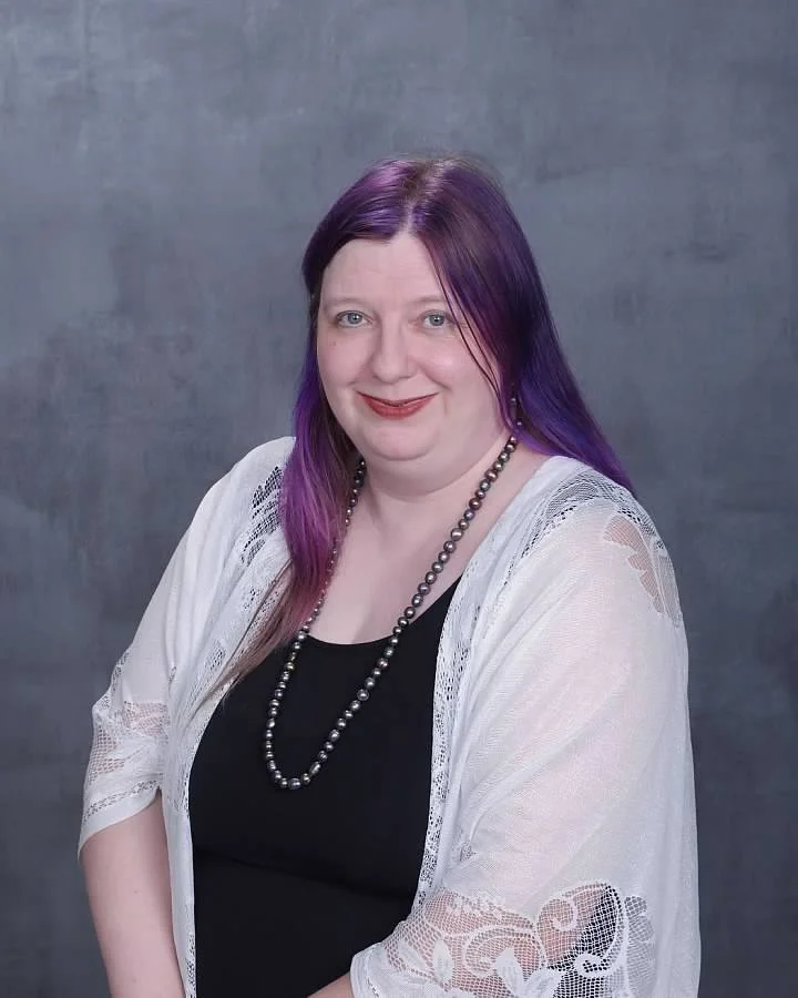 A woman with purple hair wearing a black top, a white lace cardigan, and a pearl necklace, smiling at the camera against a gray background.