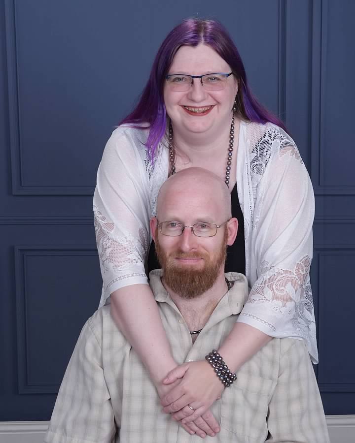 A smiling woman with purple hair, glasses, and a white lace cover-up, standing behind a man with a beard, glasses, and a beige checkered shirt, who is seated in front of a blue paneled wall.