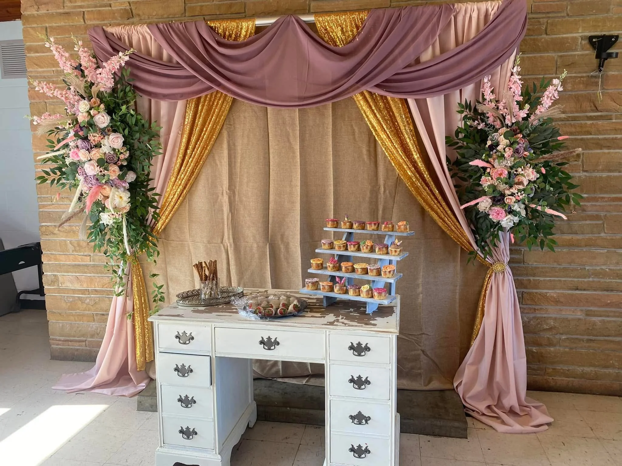 Decorative backdrop with pink, purple, and gold fabric drapes, and large floral arrangements on both sides, behind a white dresser displaying cupcakes on a tiered stand.