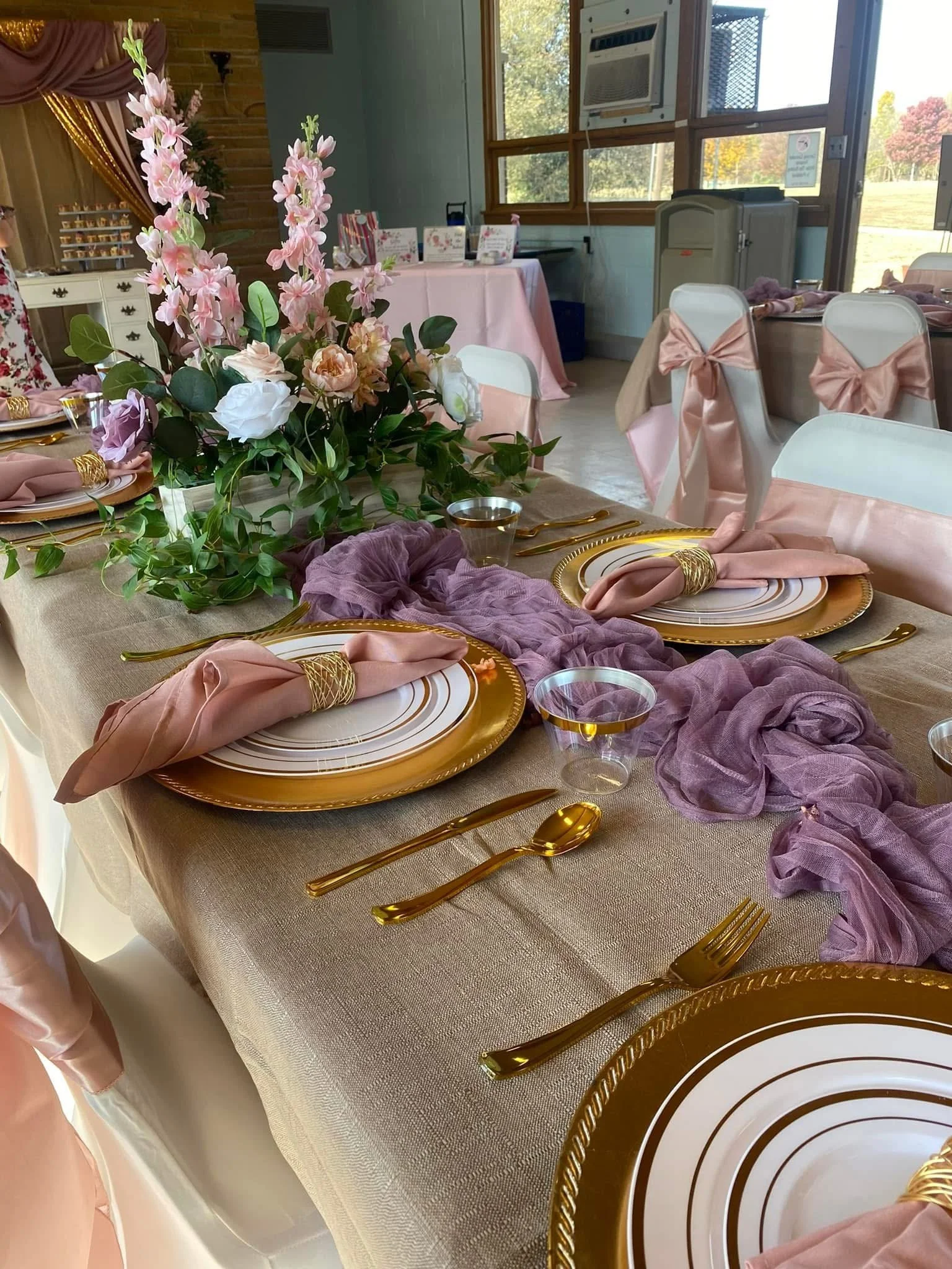A decorated banquet table with pink and gold accents, featuring floral centerpieces with pink and white flowers and green leaves, surrounded by gold-rimmed white plates, pink napkins with gold napkin rings, gold flatware, and clear plastic cups. The chairs are white with pink fabric bows on the back. In the background, there are additional tables, a pink table with signs and decorations, and a window with outdoor scenery.