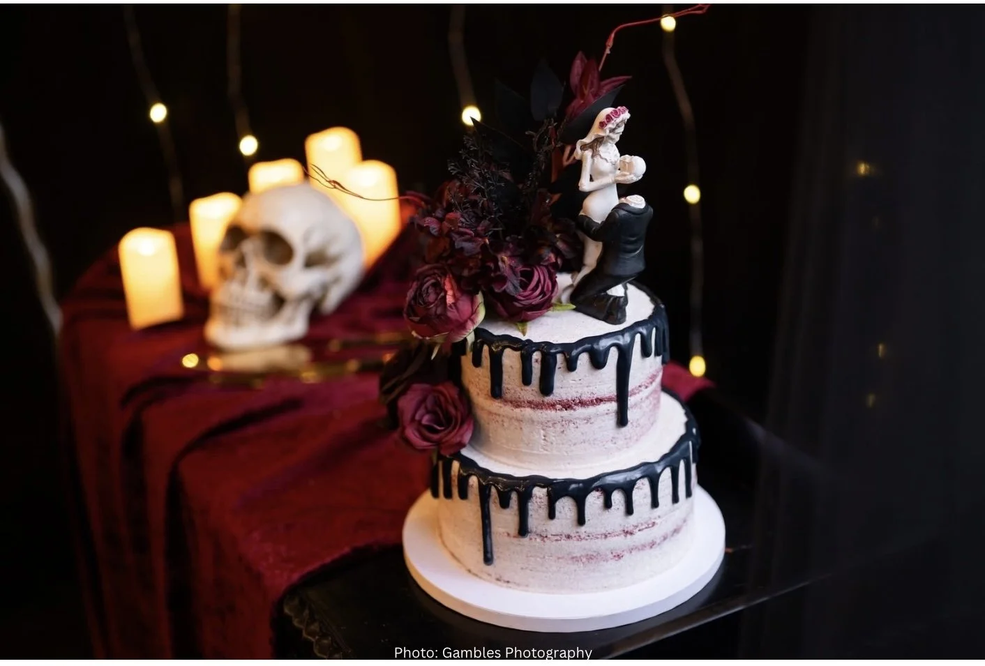 A white, red velvet, three-tiered wedding cake with black drip icing, decorated with purple roses and a bride and groom topper, set on a black table against a dark background with lit candles and skull decorations.