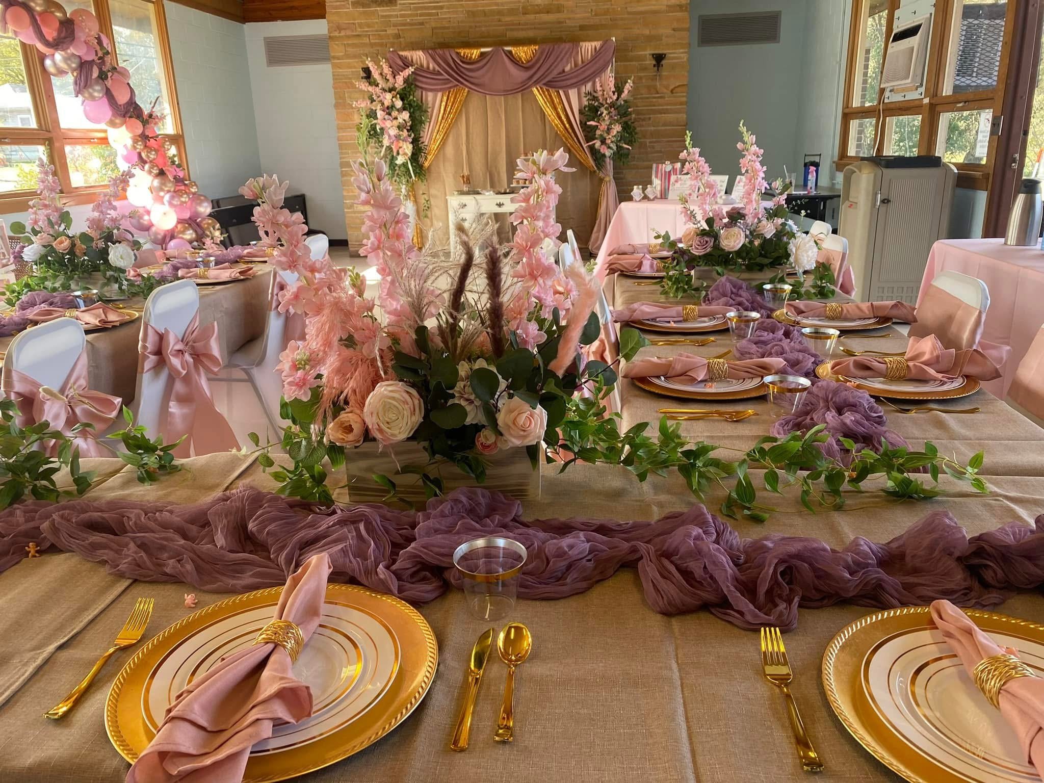 Decorated event table with pink and purple drapes, floral arrangements, and pink cloth napkins in a room with large windows and a cherrywood accent wall.