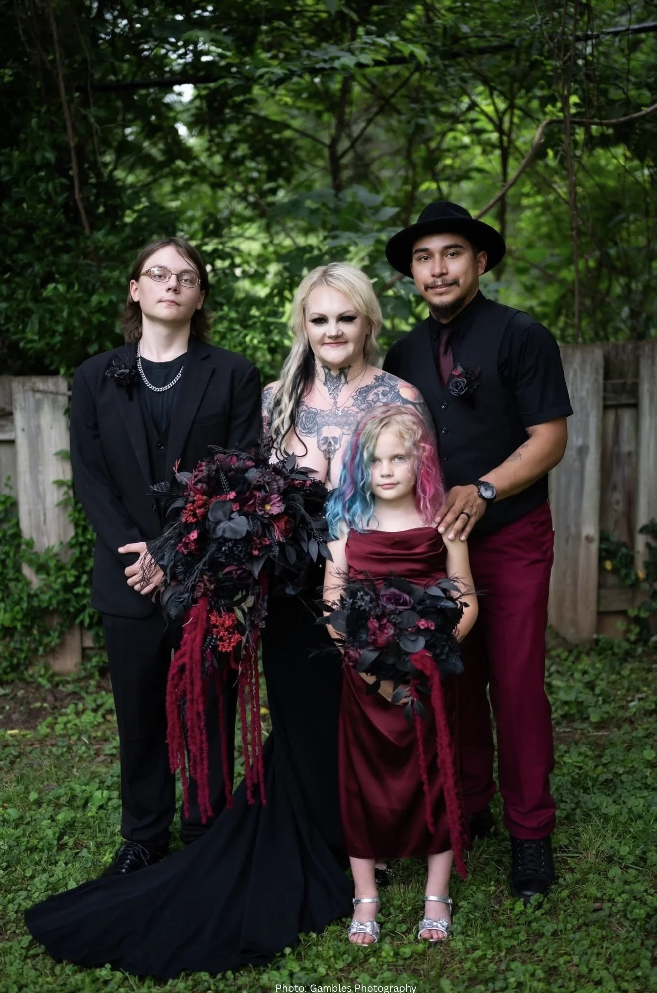 Group of four people standing outdoors in front of green trees. One young woman in black suit and glasses, one blonde woman with tattoos and dark makeup, a young girl with colorful hair in a red dress, and a man in a black hat and dark clothing.