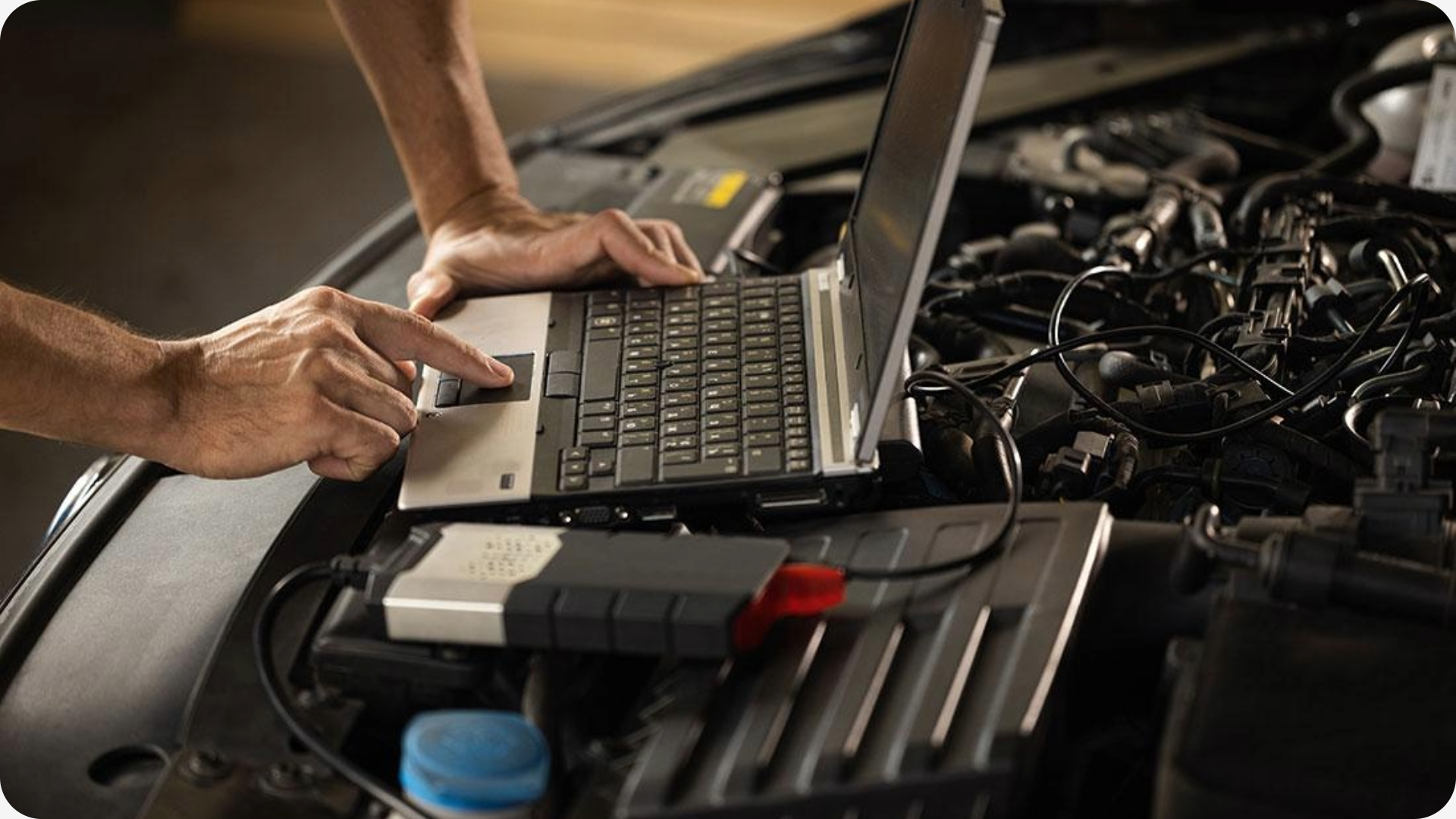 Een persoon werkt met een laptop op een open motorkap, omringd door vele auto's onderdelen en kabels.