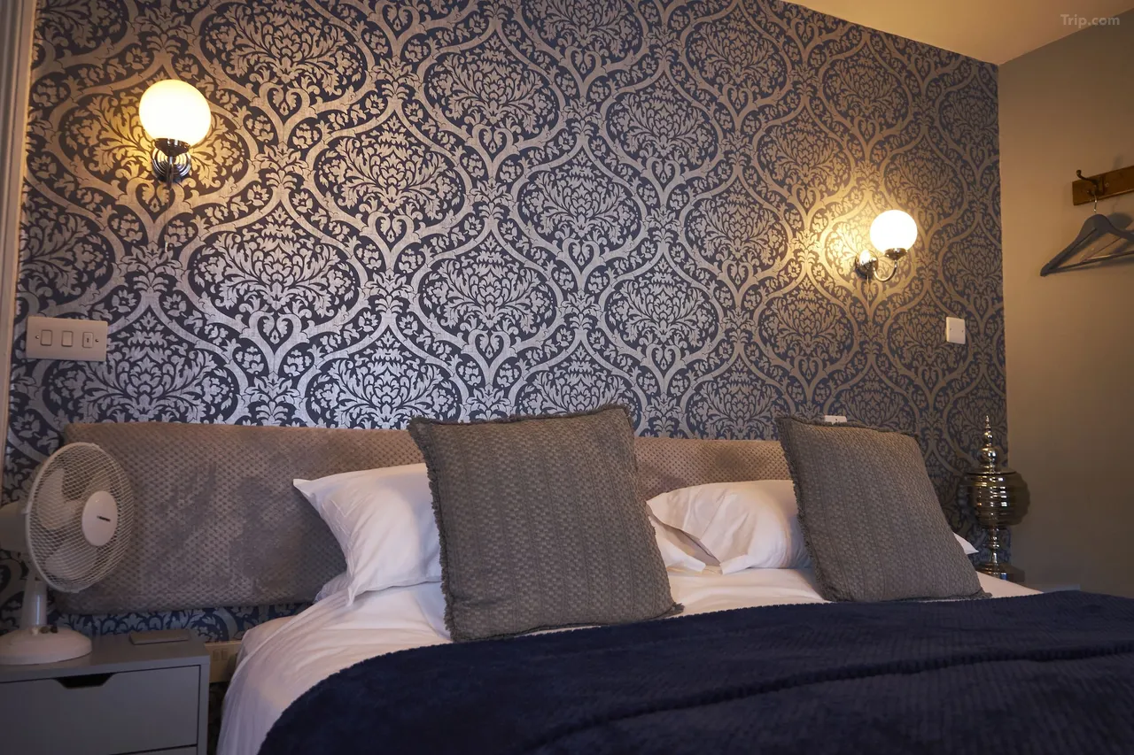 Our premium Coachmans Room at The Coachmakers Arms. A bedroom with a patterned dark and gray wallpaper, cozy bed with white sheets, grey pillow and a navy blue bedspread.