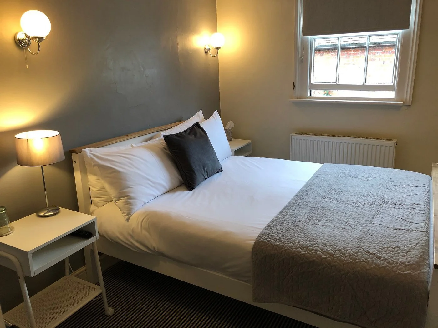 Our premium Carriage Room at The Coachmakers Arms. A cozy bedroom with a white bed, gray accent wall, nightstand with a lamp, and a window with a view of the garden outside.