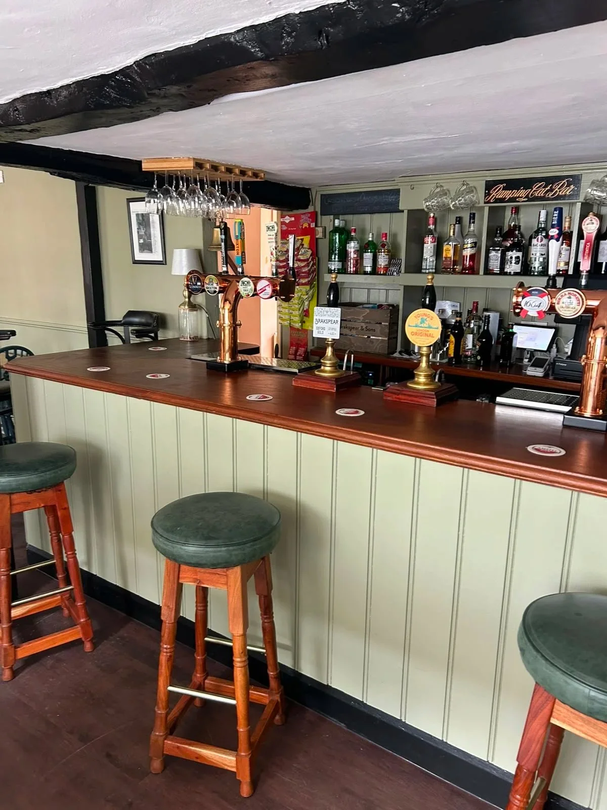 Inside at The Coachmakers arms at the main bar area with a curved wooden counter, green upholstered bar stools, and shelves stocked with liquor bottles and glasses ready for you to come and sample.