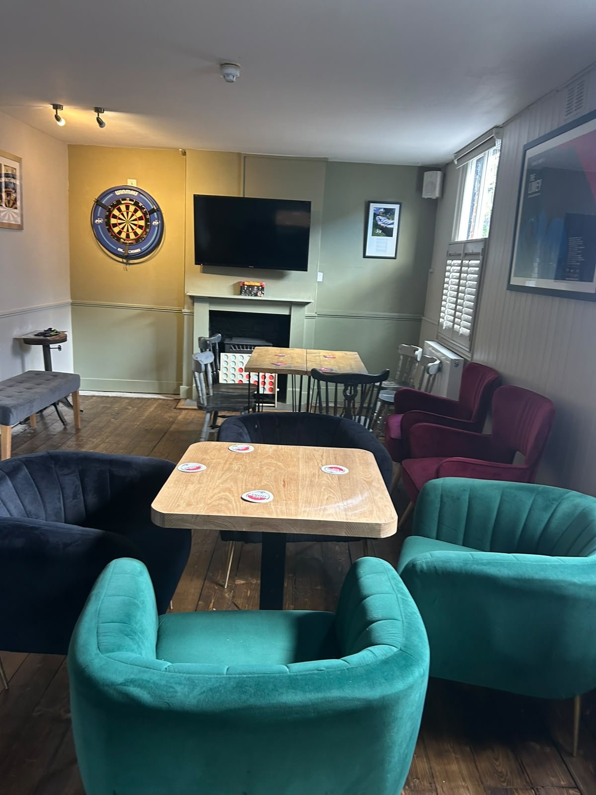 Cozy bar and lounge area inside The Coachmakers Arms with colourful chairs, tables, dartboard on the wall, television, and framed pictures.