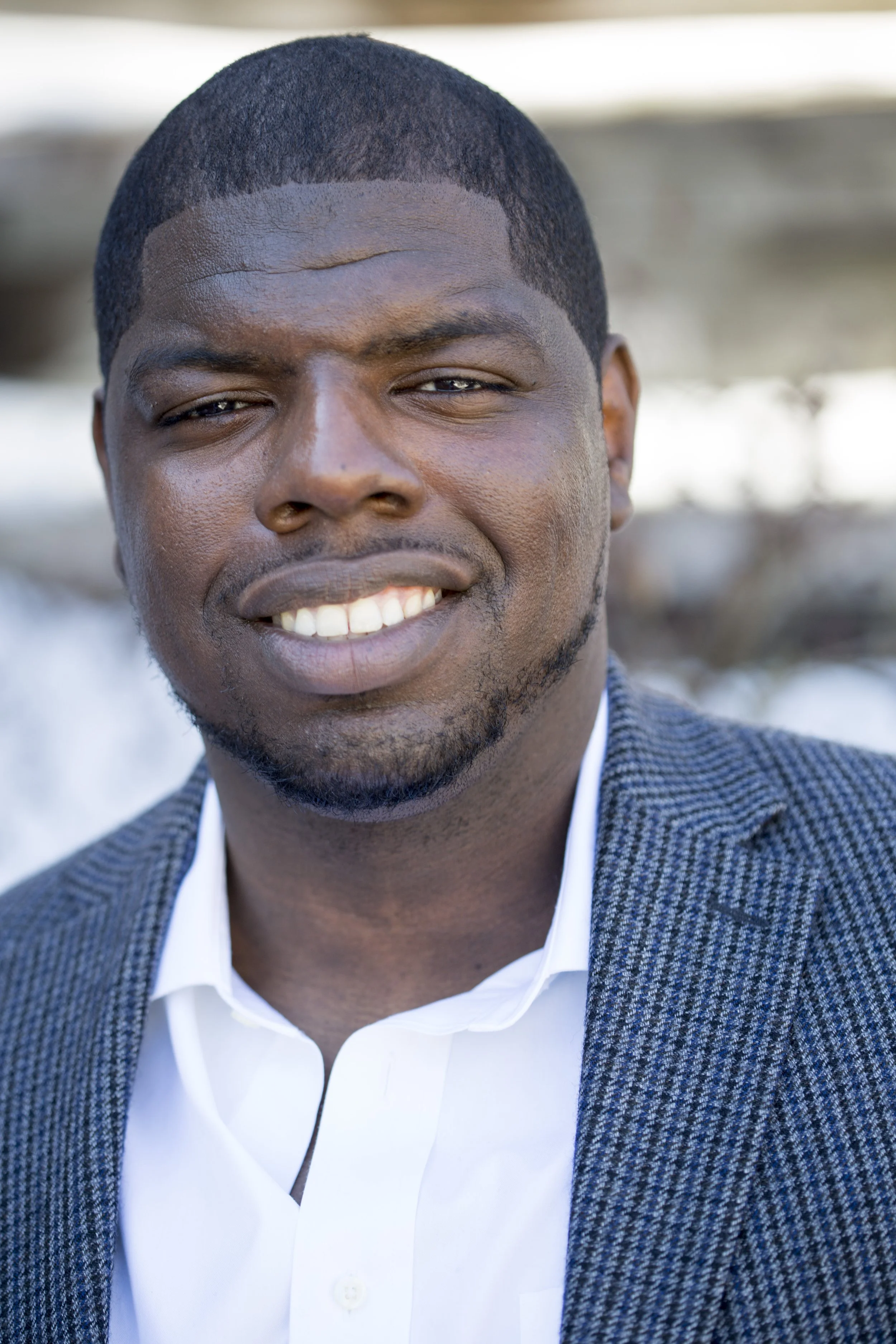 Close-up of a smiling young man with short black hair, brown skin, wearing a white shirt and a dark-colored patterned blazer. The background is blurred.