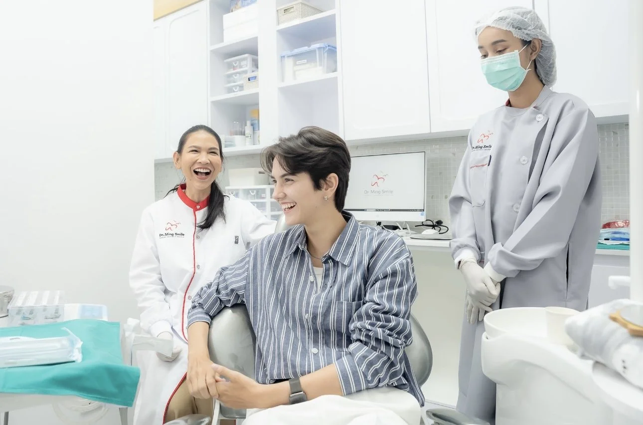 A woman is sitting in a dental chair at the dentist's office, smiling and talking with a female dental professional. Another female staff member wearing a surgical mask and gown stands nearby, also smiling in the background.