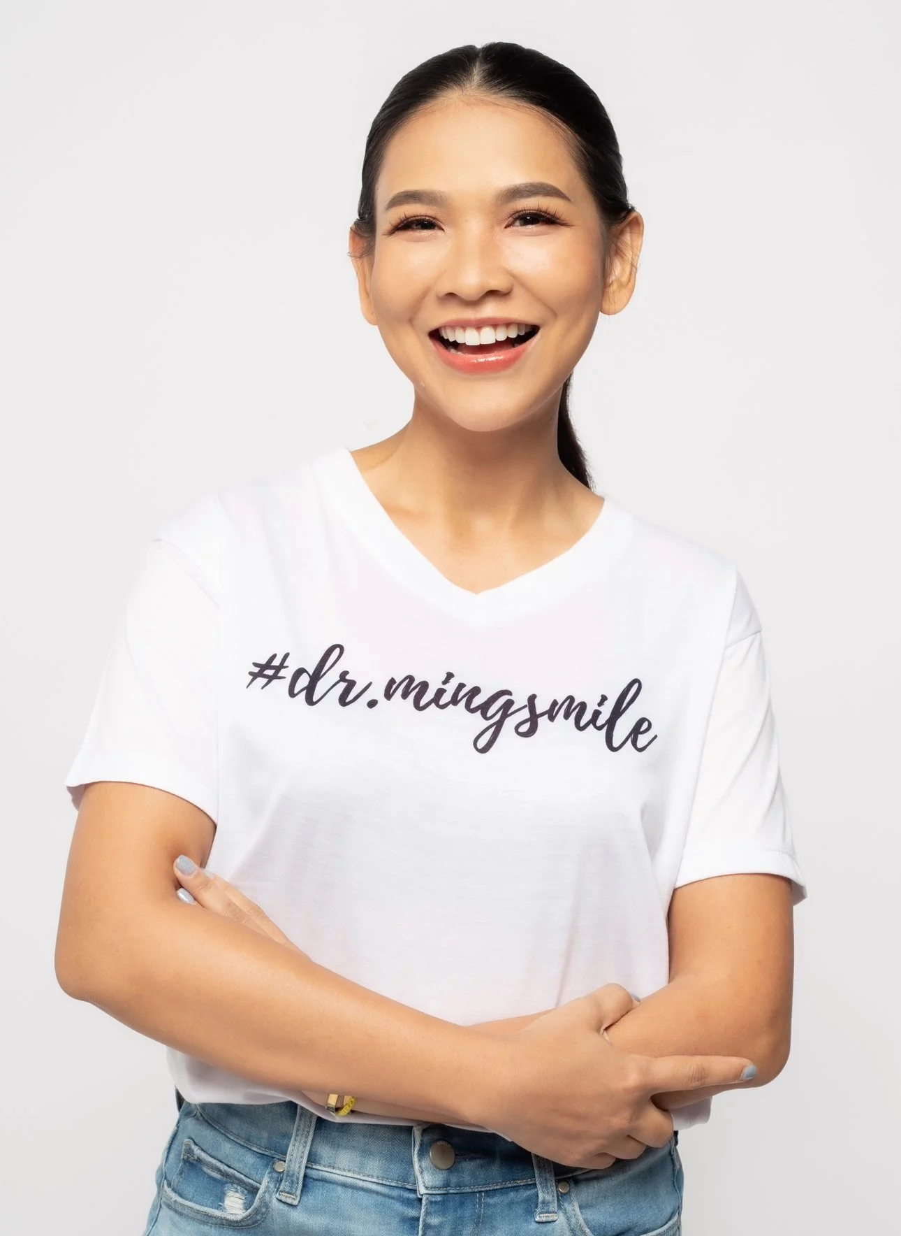 A young woman with dark hair tied back, smiling brightly with her arms crossed, wearing a white t-shirt with the hashtag '#dr.mingsmile' written on it, against a plain white background.