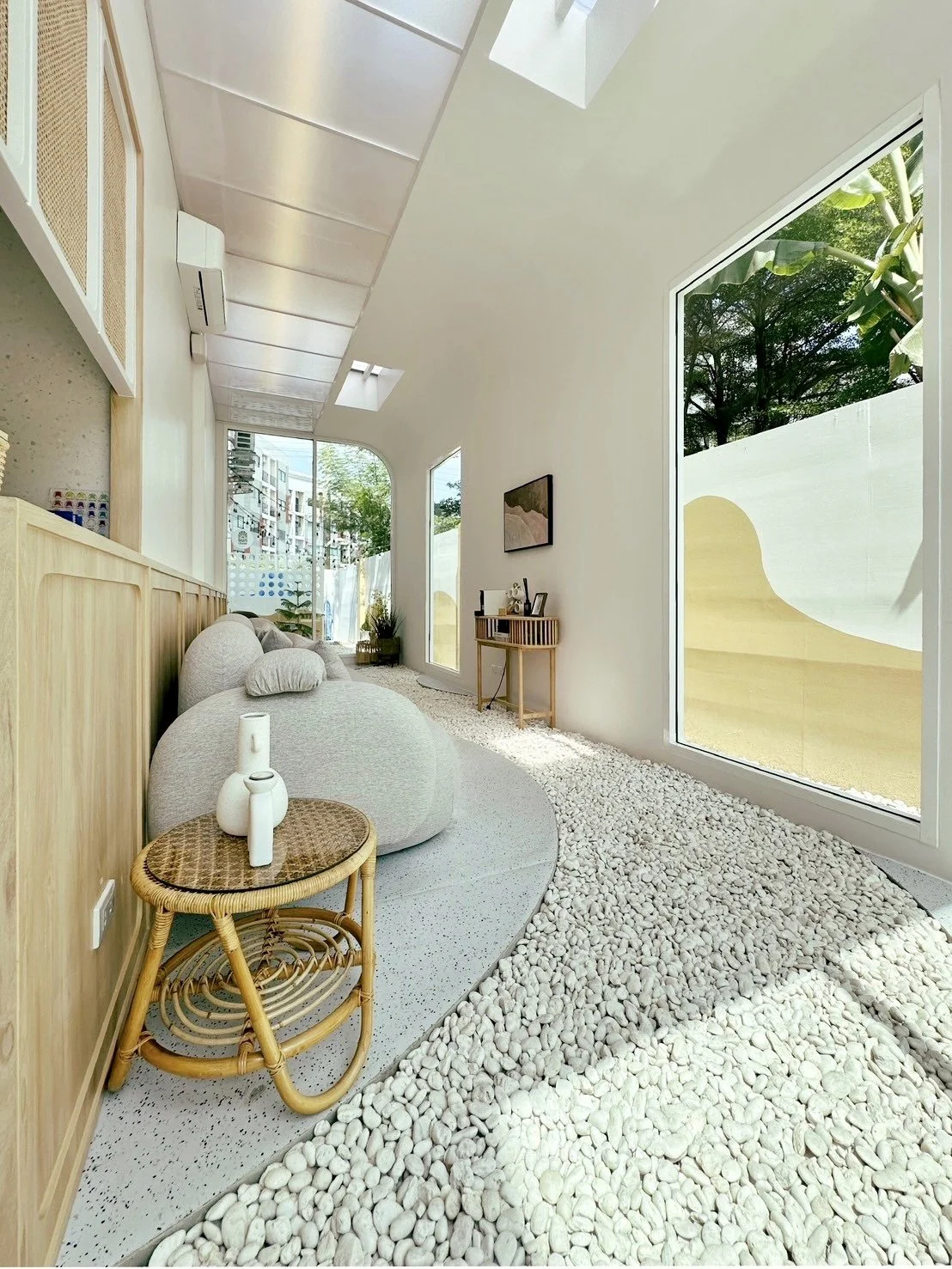 A modern indoor space with large windows, a beige sofa, a small round wooden side table with white vases, a wooden piece of furniture with decorative items, and a white pebble floor design.