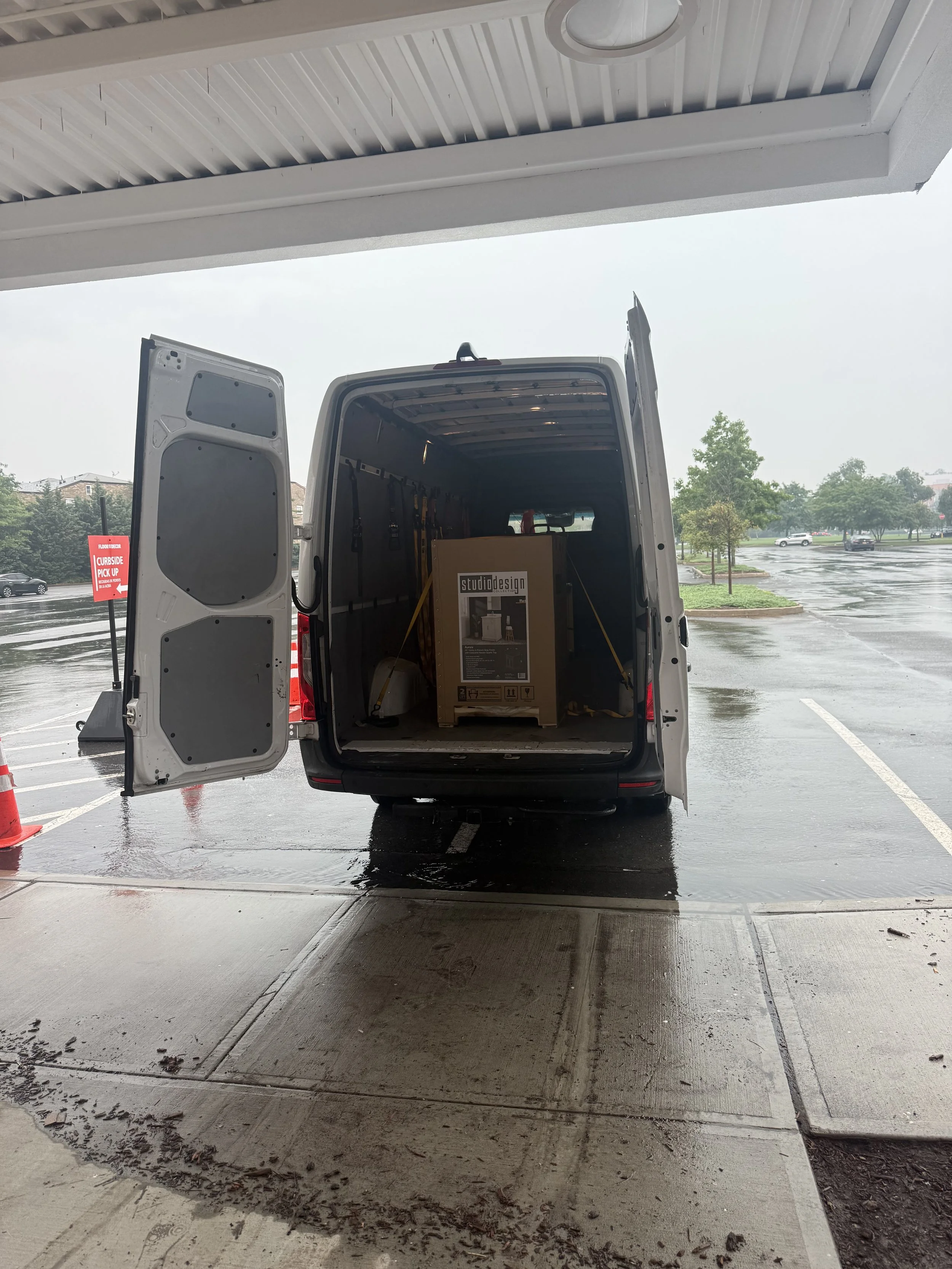 Open back of a delivery van with a large boxed item inside, parked under a building with a wet parking lot and trees in the background on a rainy day.